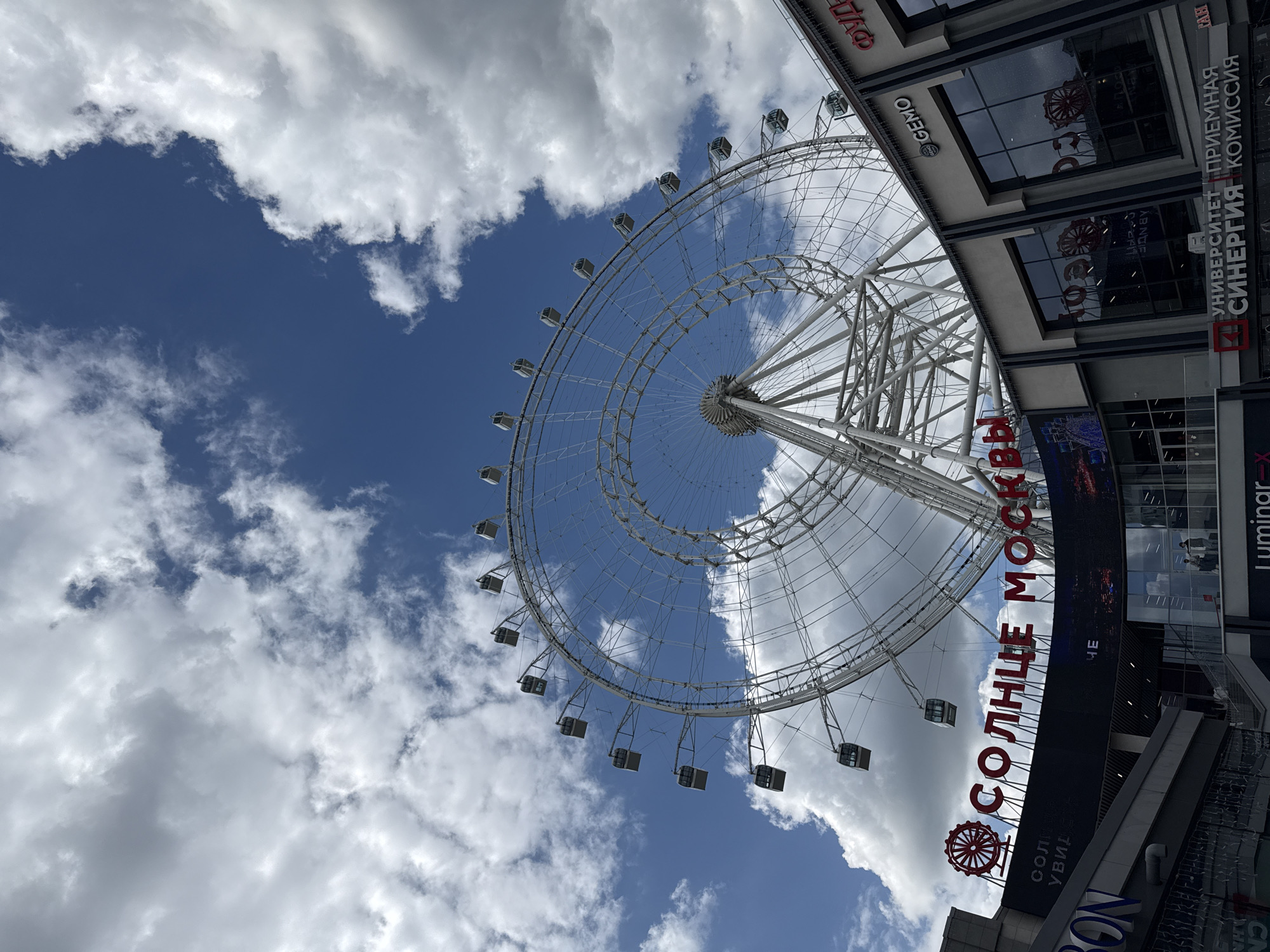 Here's a description of the image:
The image captures a low-angle view of the Ferris wheel known as "Sun of Moscow" (Солнце Москвы) in Moscow, Russia. The large, white Ferris wheel dominates the frame, contrasting with a partially cloudy blue sky. The gondolas are visible along the wheel's circumference.
A portion of the building adjacent to the Ferris wheel is in the frame, featuring advertising and signage including the word "GEMO", and "УНИВЕРСИТЕТ СИНЕРГИЯ ПРИЕМНАЯ КОМИССИЯ" (University Synergy Admission Commission), and the word "СОЛНЦЕ МОСКВЫ" in red lettering.
