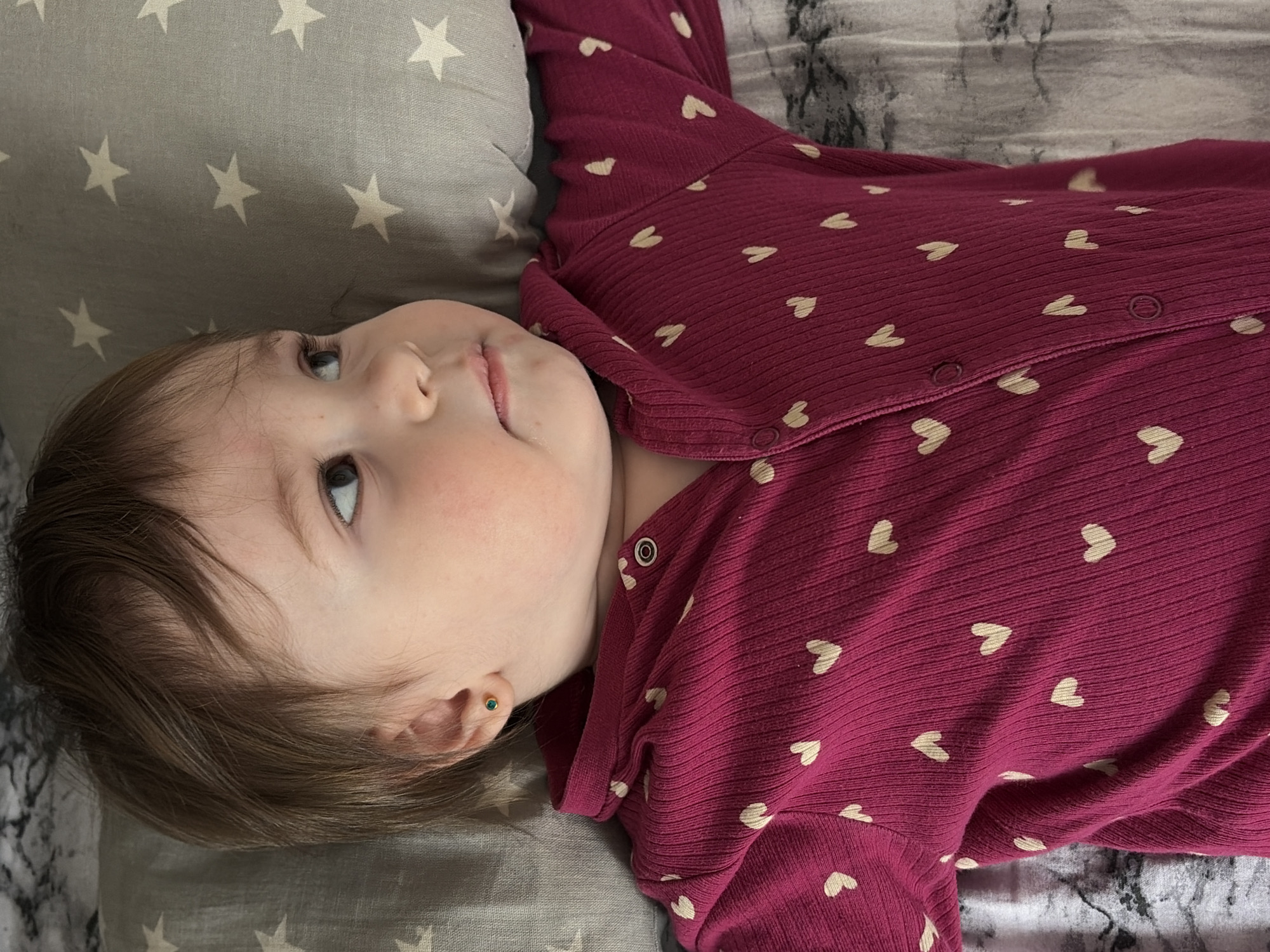 A young child is depicted lying supine on bedding in an indoor setting within Chaikovsky, Russia. The child, with light skin and short brown hair, has its head turned to the right and eyes directed upwards and towards the right. A small, round green stud earring is visible in the lobe of the child's left ear. The child is dressed in a long-sleeved, ribbed magenta garment, possibly a onesie, patterned with numerous small, light-colored heart shapes. Snap fasteners are visible down the center front of the garment. The child's head rests on a gray pillow adorned with white star patterns. A lighter, mottled gray and white fabric, indicative of a bedsheet, is visible beneath the child's body.