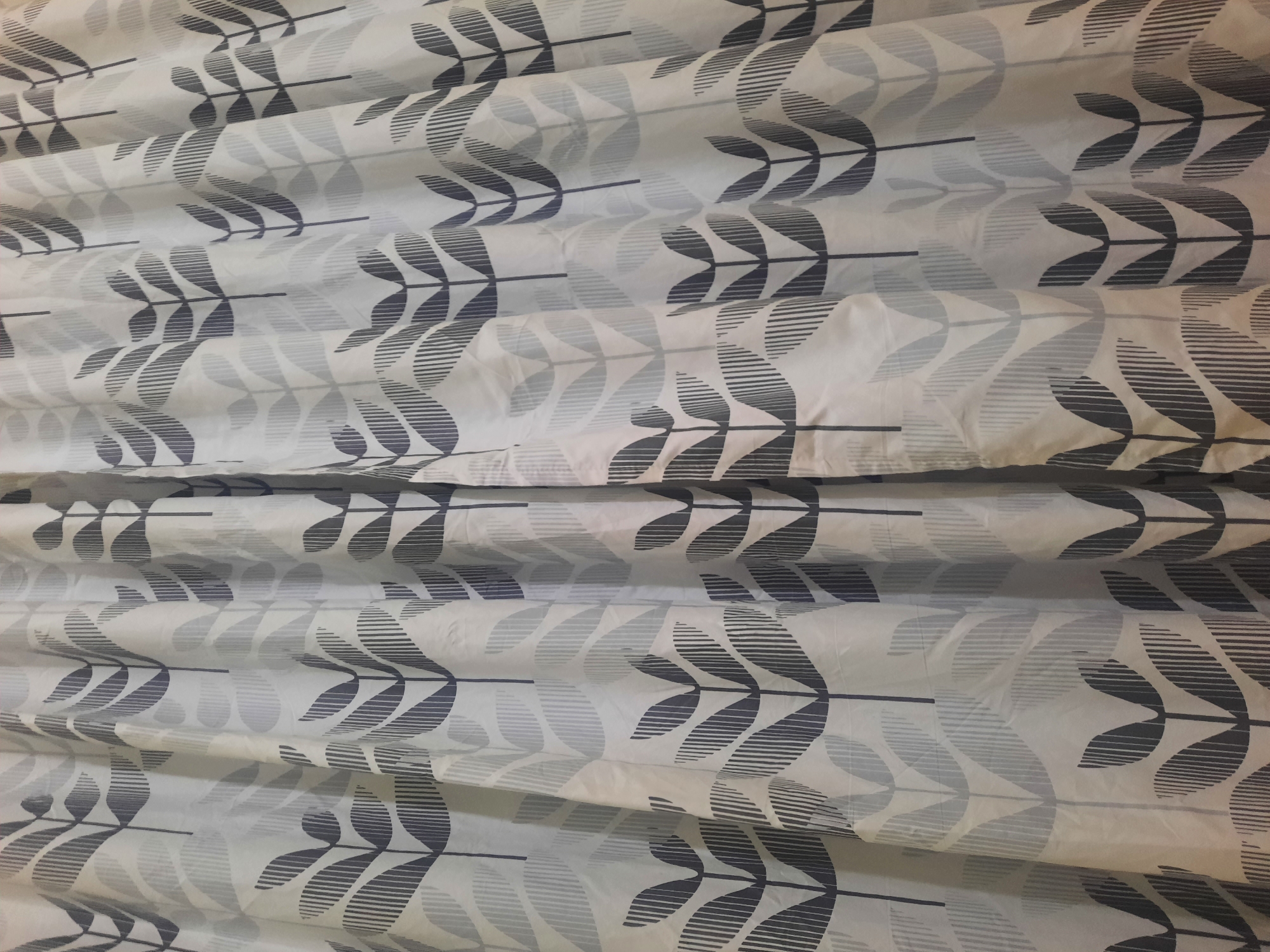 This is a close-up, eye-level, medium shot photograph of a white shower curtain with a repeating pattern of stylized dark gray and light gray leaves. The leaves are arranged in vertical rows, with each leaf resembling a sprouting plant or a cluster of petals. The texture of the fabric is visible, showing folds and creases due to the way the curtain is hung. The lighting appears to be artificial, casting subtle shadows and highlighting the folds. There are no people, animals, or other objects present in the image. The location of this shower curtain in the city of Lhokseumawe, Indonesia, cannot be determined from the visual information provided in the photograph alone.