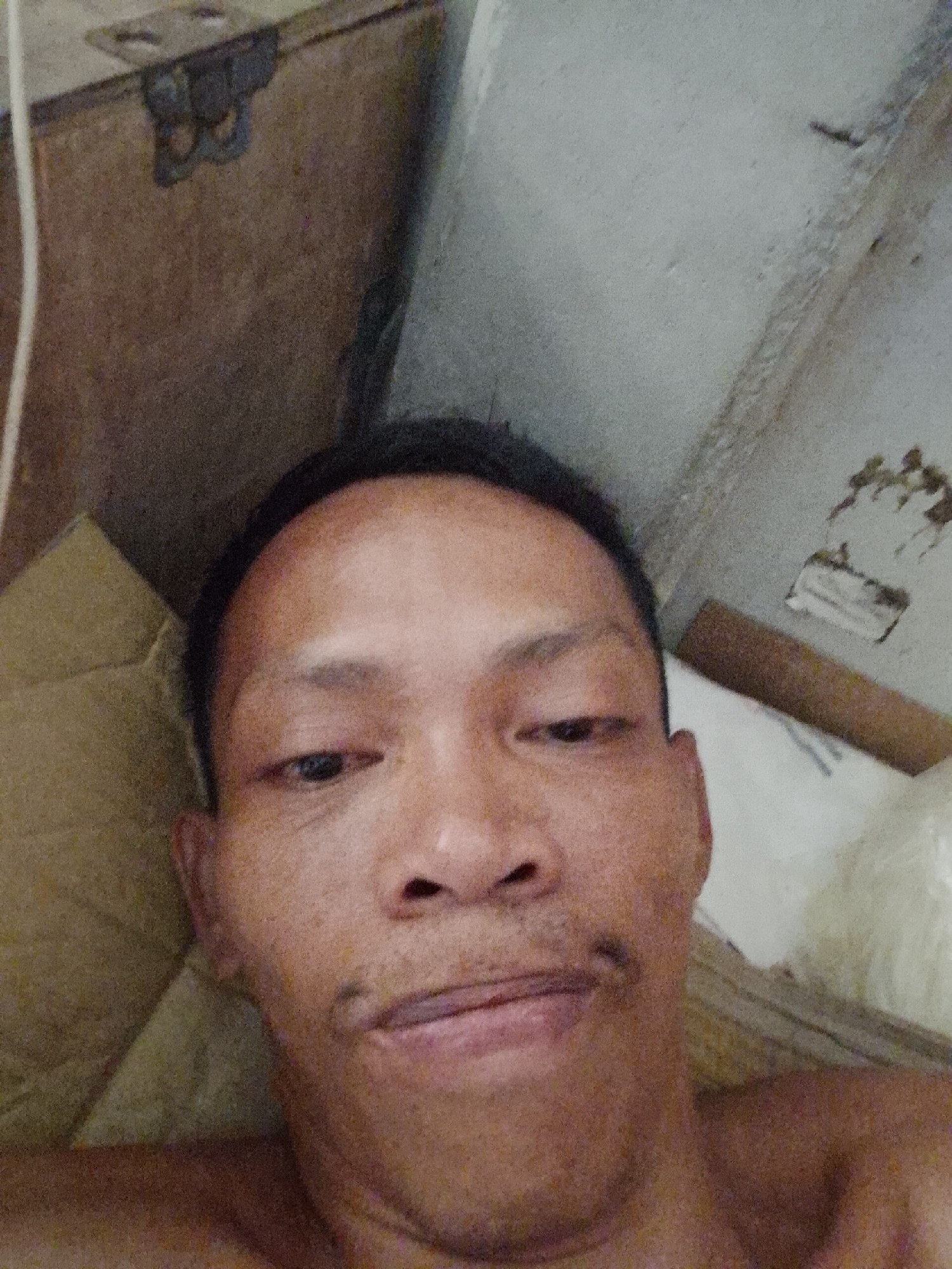 A close-up, angled selfie shot of a middle-aged Asian man with dark hair, lying down indoors. He has a neutral expression. Behind him are various objects including what appears to be a pillow, a white surface, and possibly part of a wooden box or structure with metal hardware. The location is presumed to be Bukit Ketok, Indonesia.