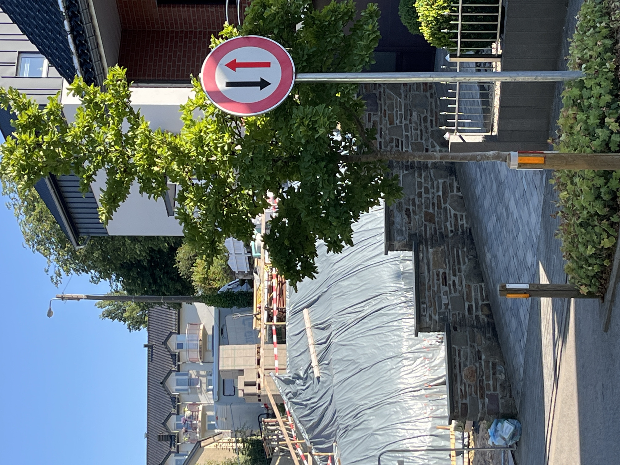 The image shows a residential street in Saint Vith, Belgium, under construction. A traffic sign indicating two-way traffic is visible in the foreground, with a tree to its left. Behind the sign, a construction site is covered with a large gray tarp. Buildings, including a house with a brick facade and a multi-story residential building, are present in the background. A street lamp is also visible. The scene is illuminated by sunlight, casting shadows on the ground.