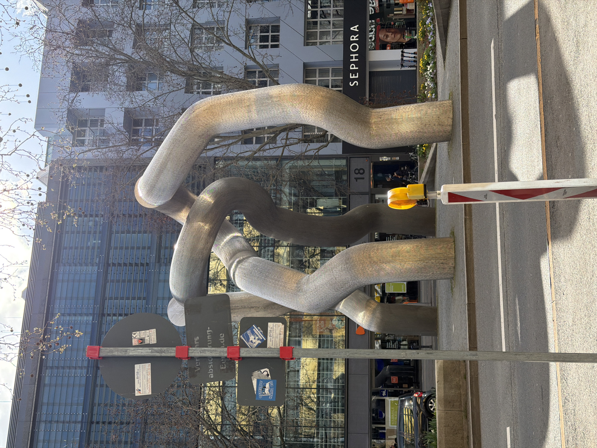 The image shows a large, intertwined sculpture made of a grey, textured material, located in Berlin, Germany. The sculpture sits on a sidewalk next to a street. Behind the sculpture is a modern building with large windows and the word "SEPHORA" visible above the entrance, which is numbered 18. A street sign is in the foreground, partially obscuring the view. A traffic barrier with a yellow light and red and white stripes is also visible. The scene is brightly lit, with strong sunlight casting shadows on the ground. The buildings reflect the light. Bare branches of trees frame the top portion of the image, indicating it is likely winter or early spring. A black car is partially visible in the background, parked along the curb. Stickers are on the road sign.