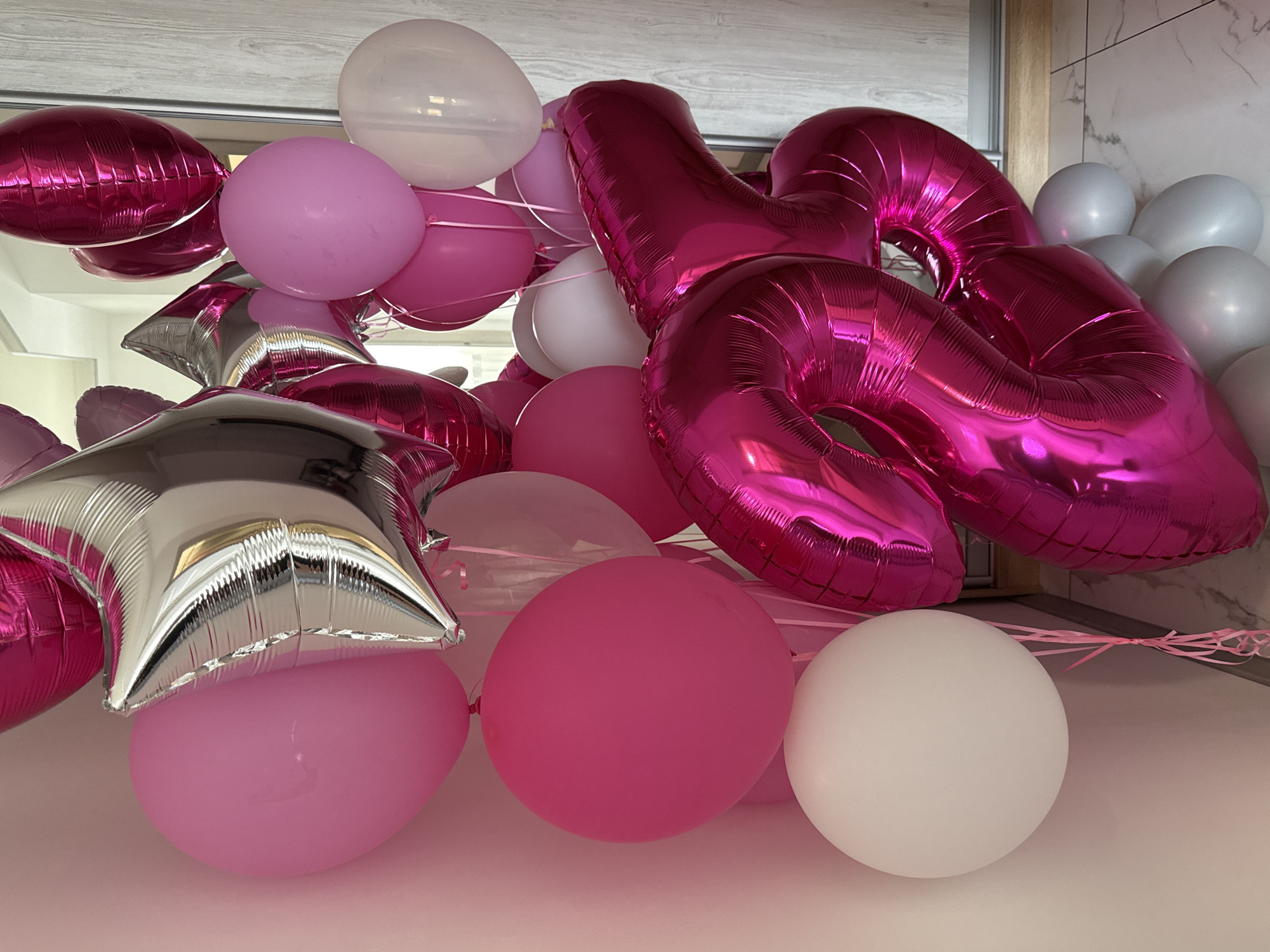 The image displays a collection of balloons in various sizes and colors, predominantly pink and silver. The balloons are arranged in a bouquet, with some larger, foil-shaped balloons forming the numbers "25" in a shiny fuchsia color. There are also several round balloons in shades of pink and white, as well as star-shaped silver balloons. The balloons are clustered together and appear to be indoors, with a pale pink wall on the left and a reflective surface, possibly a mirror or glass door, on the right showing a faint reflection of the balloons and an interior. The flooring is a light-colored tile with a subtle pattern. The overall composition suggests a celebratory arrangement, possibly for a birthday or anniversary. There are no people visible in the image, and the location is described as Unknown, Unknown.