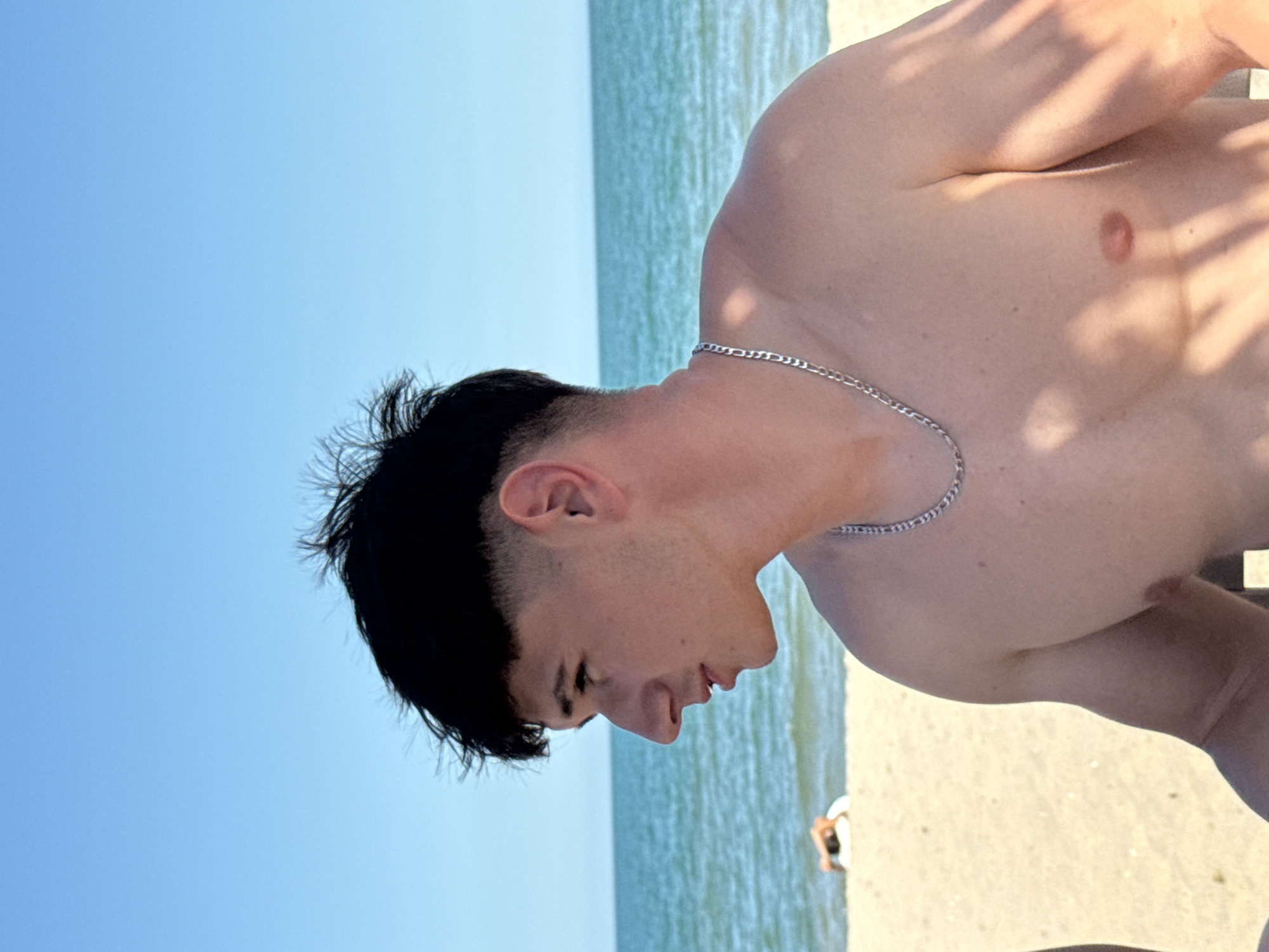 A young man with dark hair is pictured from the chest up, facing to the left. He is wearing a silver chain necklace. The background shows a beach with sand in the foreground and the ocean and sky behind. The ocean is calm and blue, and the sky is clear and bright. There are shadows cast on the man's torso, suggesting the presence of overhead objects like beach umbrellas or trees. In the far background, a person is lying down on the sand, partially obscured. The image appears to have been taken during daylight hours. The location is the beach in Varna, Bulgaria.
