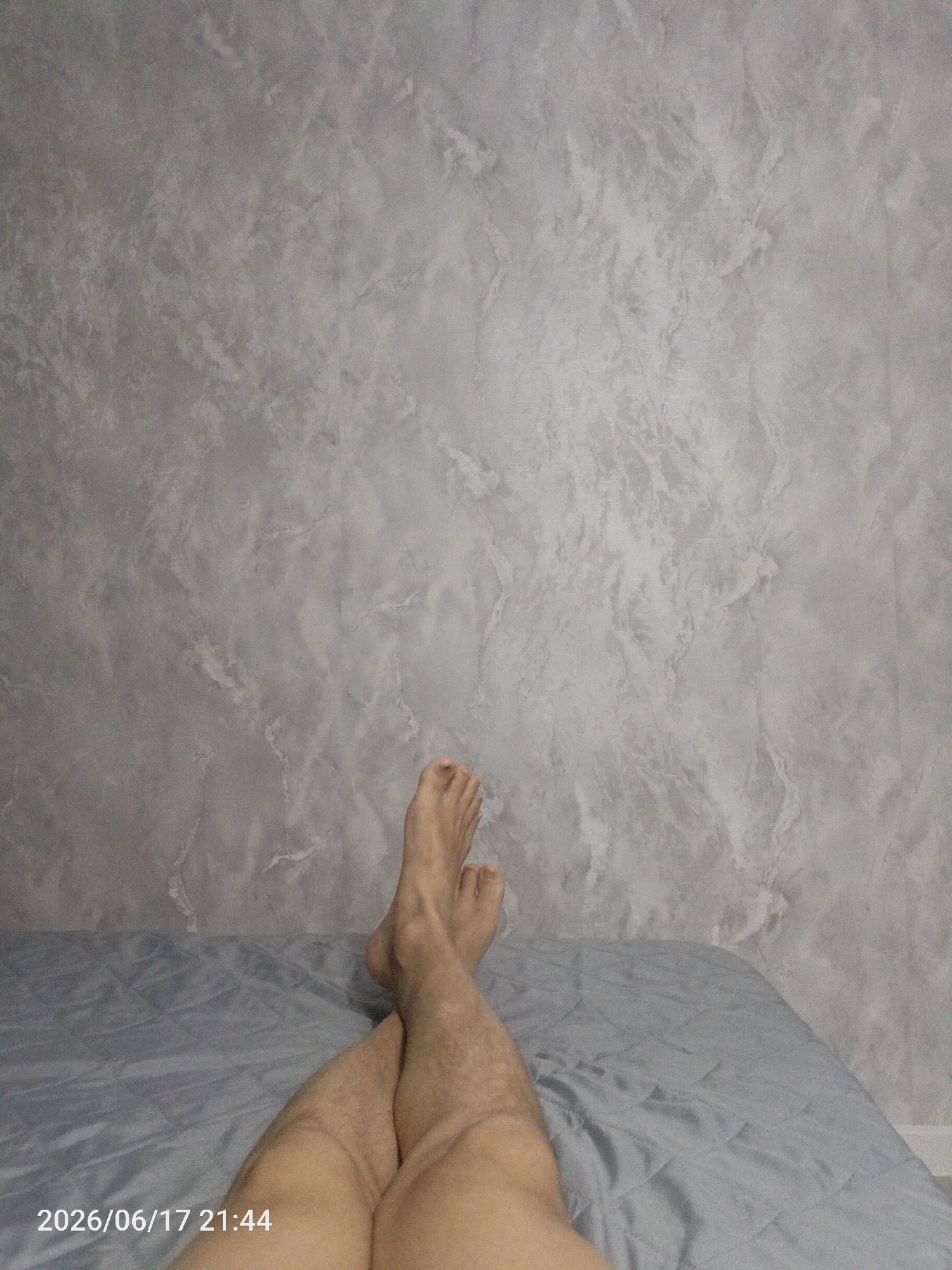 The image shows a person's legs crossed while lying on a bed. The background is a textured gray wall that resembles marble or stucco. The bed is covered with a light blue quilted comforter. The lighting suggests it is indoors, possibly at night, as indicated by the timestamp "2026/06/17 21:44" visible in the lower-left corner of the image. The scene is casual and relaxed, with the person appearing to be at ease. There are no specific clues to suggest the weather or a particular location within Pavlodar, Kazakhstan, beyond the general domestic setting. The focus is on the legs and the simple environment.