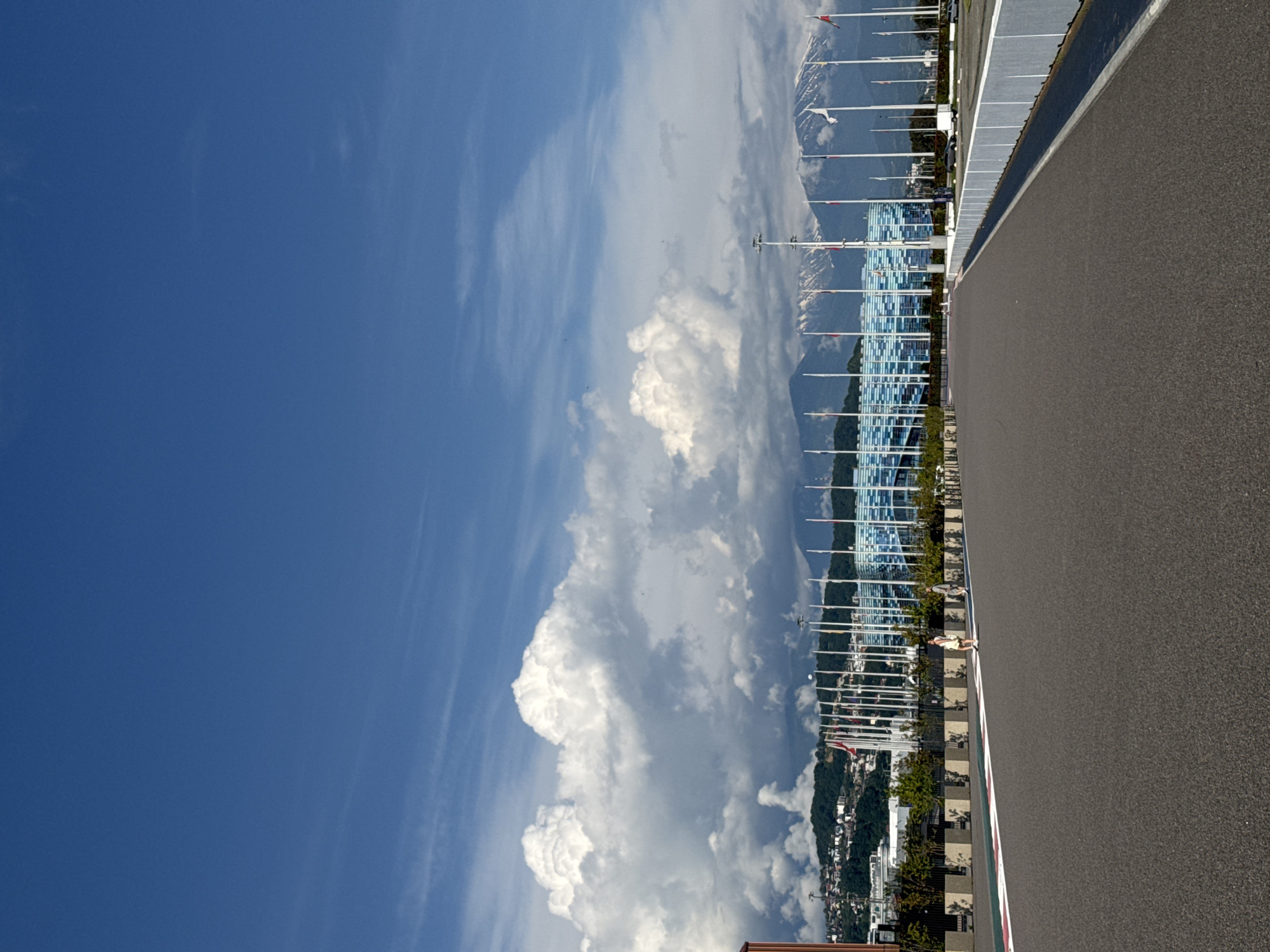 The image displays an outdoor scene within the city of Sirius, Russia, featuring an open asphalt track with architectural structures and natural landscapes.
The upper portion of the frame shows a vast blue sky, predominantly clear but with significant formations of large, white cumulus clouds concentrated across the left and central mid-sky. Lighter, wispy clouds are also present higher in the atmosphere.
In the distant background, snow-capped mountains are visible, their peaks partially veiled by clouds. Closer to the mid-ground, a series of green, tree-covered hills ascend, dotted with numerous residential or commercial buildings.
A large, modern building with a distinctive blue and white grid-patterned facade dominates the central mid-ground, suggesting a stadium or major venue within an Olympic park complex. In front of this building and extending across the middle distance, numerous tall, white flagpoles are arrayed, some bearing flags that are not clearly identifiable. Barriers and a line of low-lying vegetation separate the track area from these structures.
The foreground consists of a wide asphalt surface, typical of a racetrack, which extends from the bottom of the frame towards the horizon. A white line borders a red and white striped curb along the left edge of the asphalt. On the right side of the track, a low-height grey and white barrier or wall runs parallel to the track, also receding into the distance.
Two individuals are visible on the asphalt surface in the mid-foreground. One person, dressed in light-colored attire, walks away from the viewer on the left side of the track. A second individual, wearing darker clothing, follows at a greater distance behind and slightly to the right of the first person. Both appear to be walking in the same direction, towards the background structures. The scene is brightly lit by natural daylight.