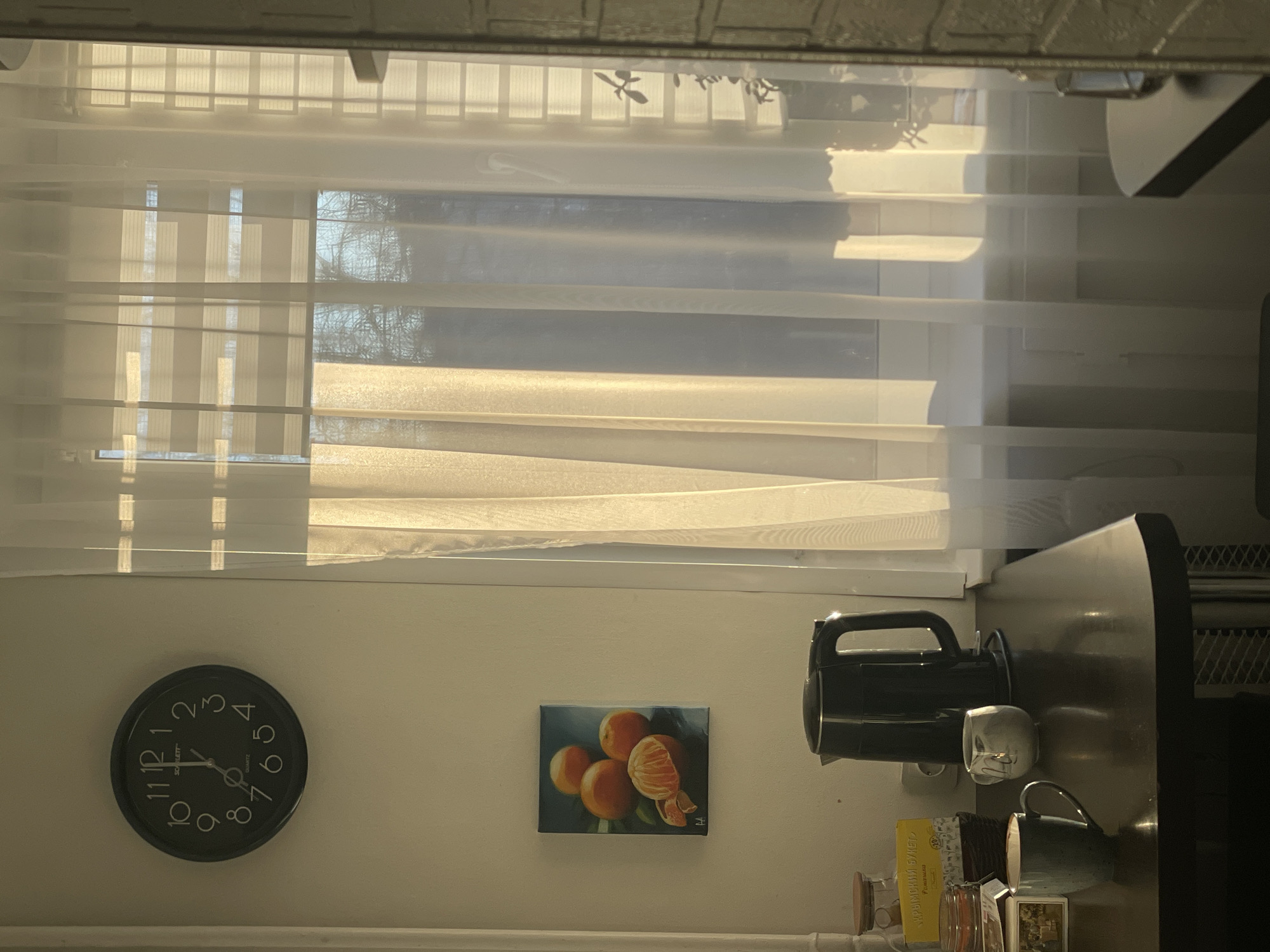 The image shows an interior scene, likely a kitchen, with a window occupying the top portion of the frame, covered by translucent blinds or curtains. The window allows natural light to illuminate the room. Below the window, the wall features a clock on the left, displaying the time as approximately 7:08. To the right of the clock, a painting of oranges is hung on the wall. Further to the right, there's a coffee maker and some mugs, along with a small collection of jars and containers on a shelf. The brand "КРЫМСКИЙ БУКЕТ" is visible on a yellow box. Based on the text in Cyrillic, the location could be Chaikovsky, Russia.