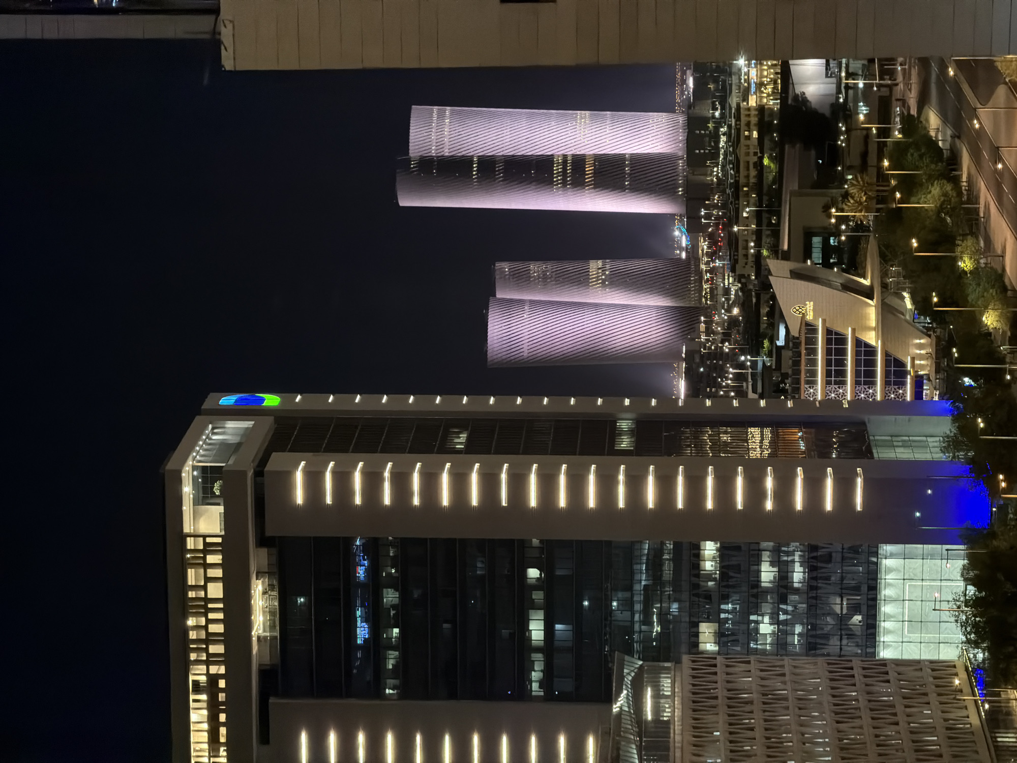 This is a nighttime exterior, eye-level, medium shot of a modern cityscape in an unknown city. In the foreground, on the left, is the side of a tall, modern building with a glass facade and vertical white lighting elements. To its right are two more buildings, one appearing to be a lower-rise commercial building with a distinct angular design and lit windows, and further back, the distinctive spiraling towers of a skyscraper complex are illuminated in pale purple. The background is dark, indicating nighttime, with numerous streetlights and building lights creating a glow across the city. Trees and streetlights line a road in the mid-ground. The overall scene depicts a well-lit, contemporary urban environment at night.