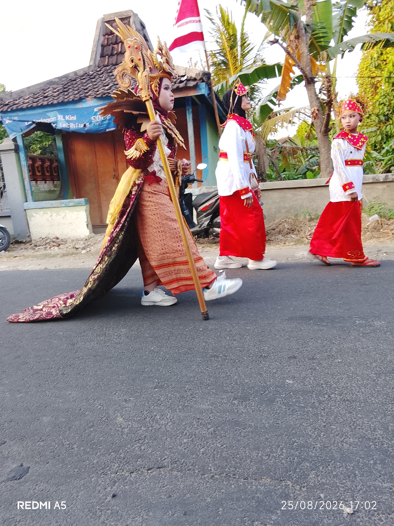 Three individuals in traditional attire are walking on a paved road in Semanding, Indonesia. The person in the foreground is wearing a large, ornate golden headdress and a maroon and gold costume with a long train, holding a wooden staff. To their left is a blue banner with white text. Behind them, two younger individuals are dressed in white shirts with red collars and trim, and long red skirts, both wearing red headbands. Trees and a building with a tiled roof are visible in the background. The image is dated 25/08/2026, 17:02.
