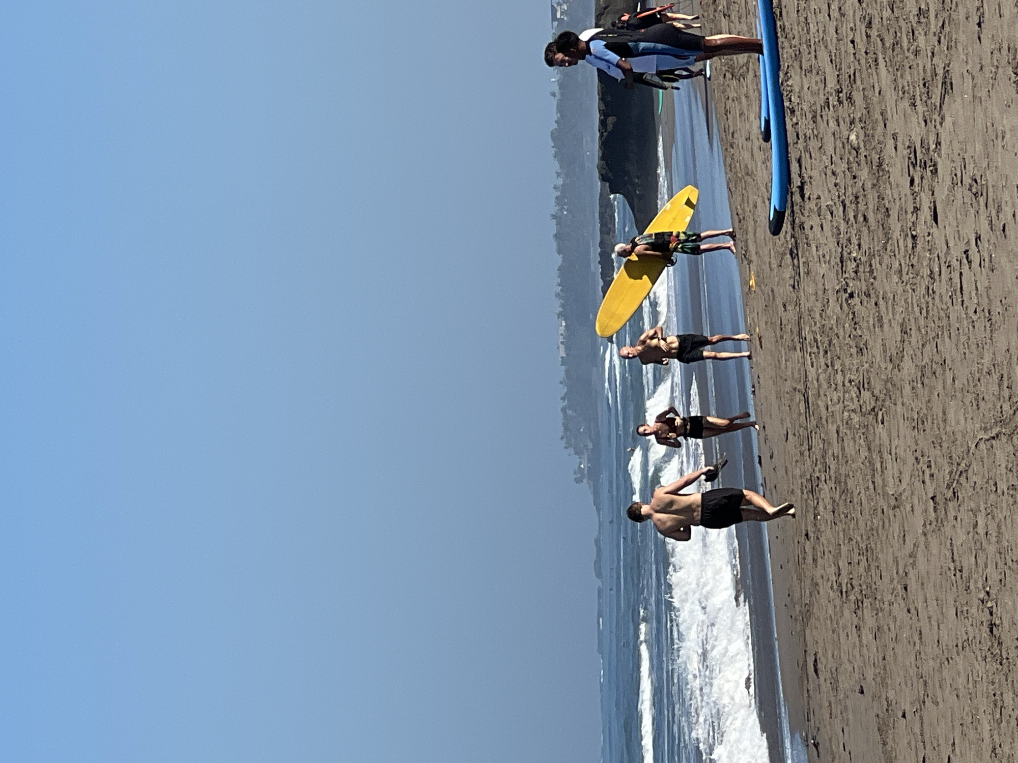 In North Kuta, Indonesia, on a sandy beach with gentle waves lapping the shore, a group of people are engaged in various activities under a clear blue sky. In the foreground, a man with dark hair is running away from the camera, wearing black shorts and carrying a dark object in his right hand. To his right, a woman in dark athletic wear is also running. Further to the right, a man with light hair is holding a bright yellow surfboard upright. Behind him, two individuals in wetsuits are standing, one wearing a blue and white wetsuit and the other a black and white wetsuit with a red trim. Two blue surfboards are lying on the sand in the lower right corner of the image. In the background, rocky outcrops jut out into the sea, and a treeline with buildings is visible on the distant horizon. The ocean is a deep blue, with white foam cresting the waves.