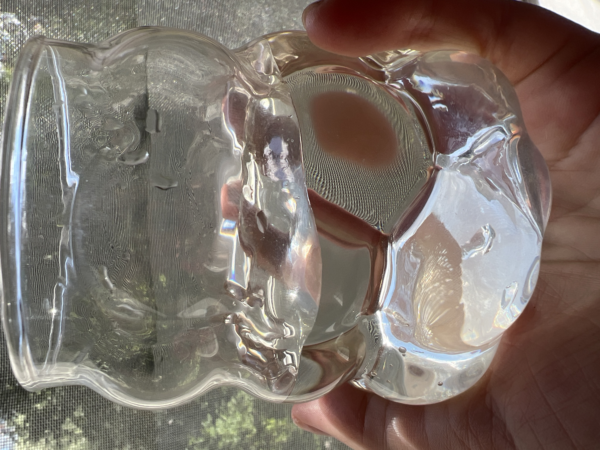 The media file features a close-up shot of a person's hand holding a clear, glass object. The object has an unusual, almost bulbous shape with a scalloped edge at the bottom. It appears to be transparent, allowing light to refract and create colorful glints and reflections, particularly on the textured surface. The glass itself has internal patterns and some small air bubbles, suggesting it might be hand-blown or a decorative piece. The background consists of a window screen and a blurred view of green foliage, indicating an outdoor setting, possibly a residential area. Sunlight streams through the window, illuminating the glass object and casting shadows. The person's hand, with visible skin texture, is holding the object firmly, suggesting an interaction with it. The overall mood is casual and perhaps contemplative, due to the focused attention on the glass and the light play. The image has been taken during daylight hours, as suggested by the bright sunlight. No visible text is present in the media file. The location context is provided as Sinyavino, Russia.