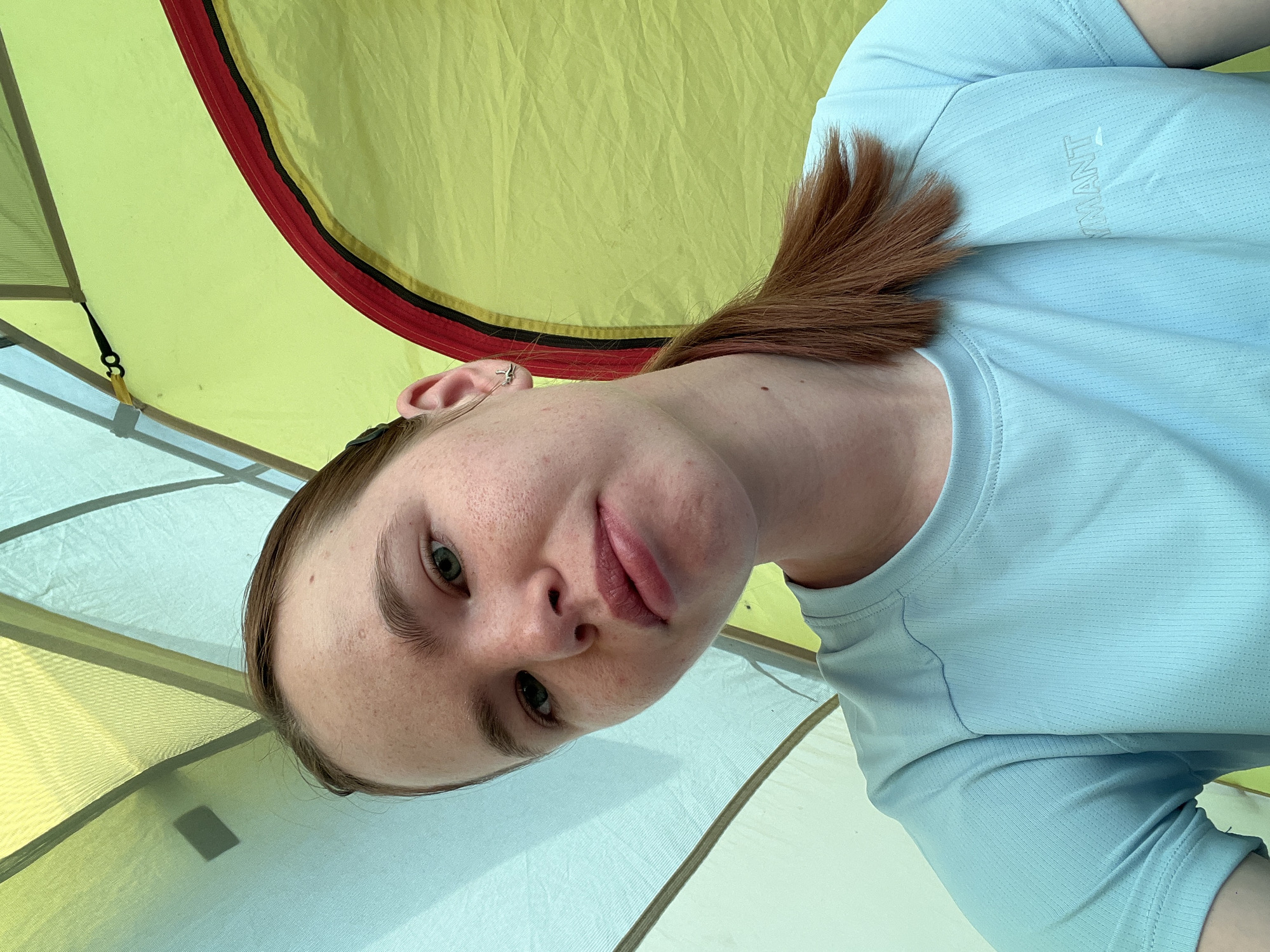 A close-up, selfie-style photograph shows a young woman inside a tent. The woman has fair skin, freckles, and reddish-brown hair pulled back in a ponytail. She is wearing a light blue athletic shirt. The tent interior features a yellow and pale green color scheme with visible seams and support structures. The photograph is taken at an angle, obscuring a clear view of the tent’s surroundings. There is no verifiable information indicating a specific location, such as Khvalynsk, Russia.