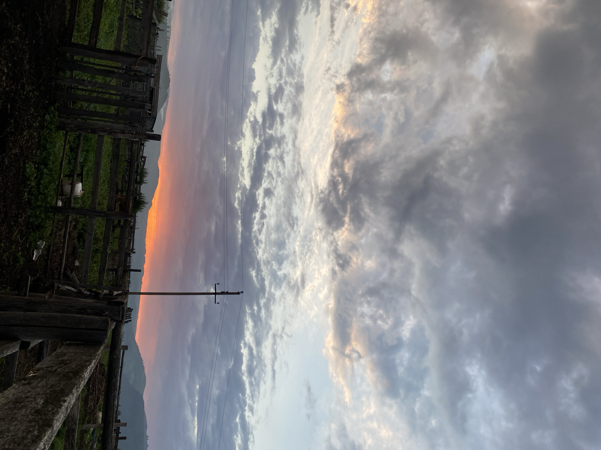 This image depicts a rural scene at dusk in Maloarslangulovo, Russia. A wooden fence encloses a muddy area in the foreground, transitioning to a grassy enclosure with more wooden fences further back. Beyond the enclosures, rolling hills are visible under a dramatic sky filled with dark, textured clouds. The horizon is illuminated by a vibrant sunset, casting a warm orange and pink glow. A solitary utility pole with wires stands to the right of center in the mid-ground. There are no people or animals clearly identifiable in the image. The overall atmosphere suggests the end of a day in a pastoral setting.