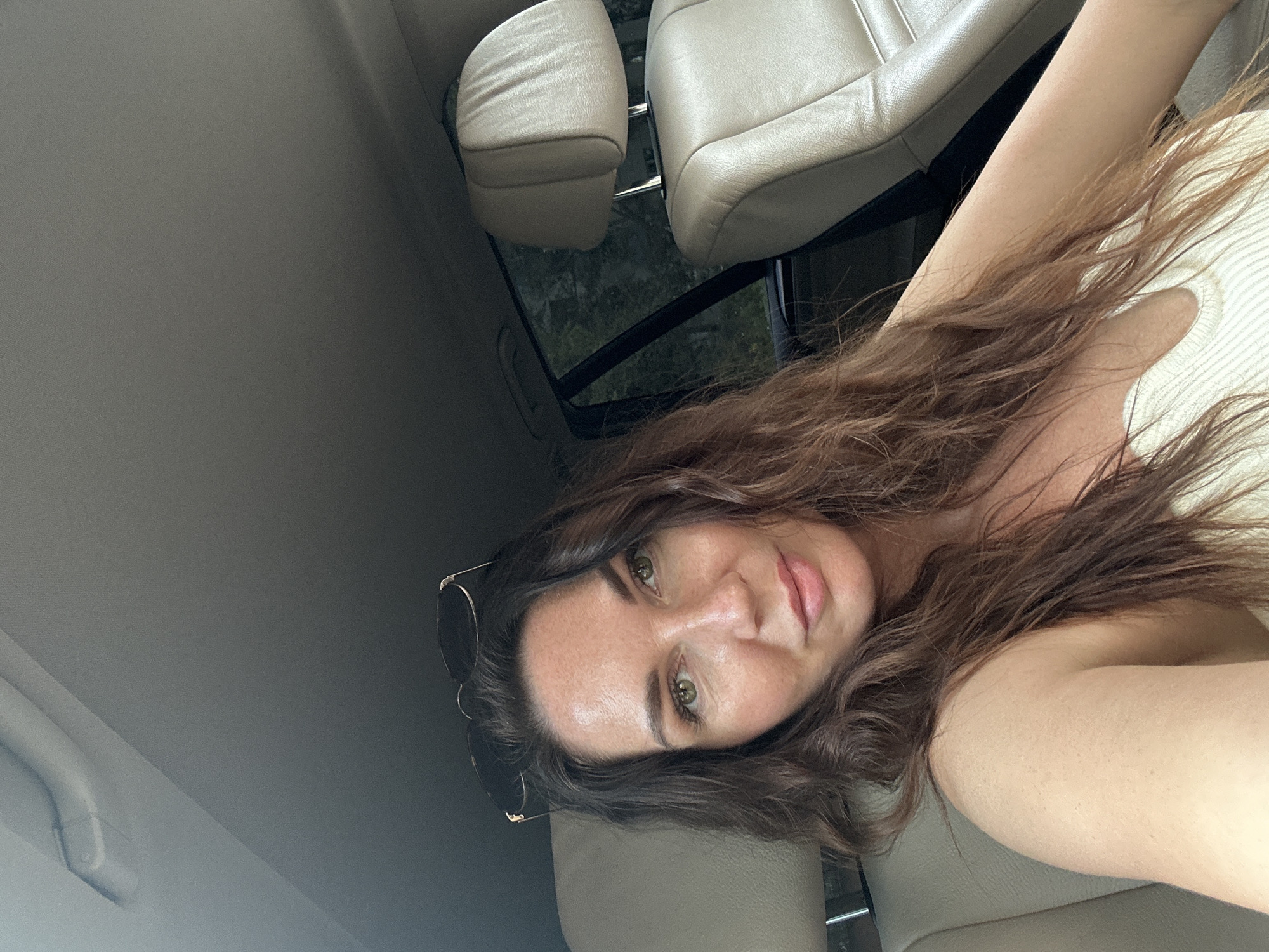 A woman with long, wavy brown hair and light green eyes is sitting in the passenger seat of a car. She is wearing a white, ribbed tank top and has sunglasses perched on top of her head. The interior of the car appears to be leather, with beige seats visible. Through the car window, trees and foliage are visible, suggesting an outdoor, possibly rural or suburban, setting. There is no specific indication of the city of Chaikovsky, Russia, in the image.