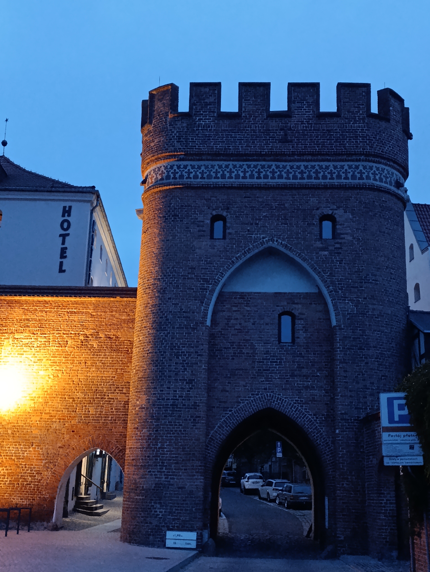 The image shows the Bridge Gate (Brama Mostowa) in Toruń, Poland, at dusk. The gate is a brick medieval structure with crenellations at the top and a large arched passage. To the left of the gate, a section of a brick wall is illuminated by a warm, yellow light, and adjacent to it is a modern white building with the word "HOTEL" printed vertically in black letters. Several cars are visible driving through the arched passage of the gate. A blue parking sign with Polish text is attached to the brick wall to the right of the gate. The sky is a deep blue, indicating the evening twilight.