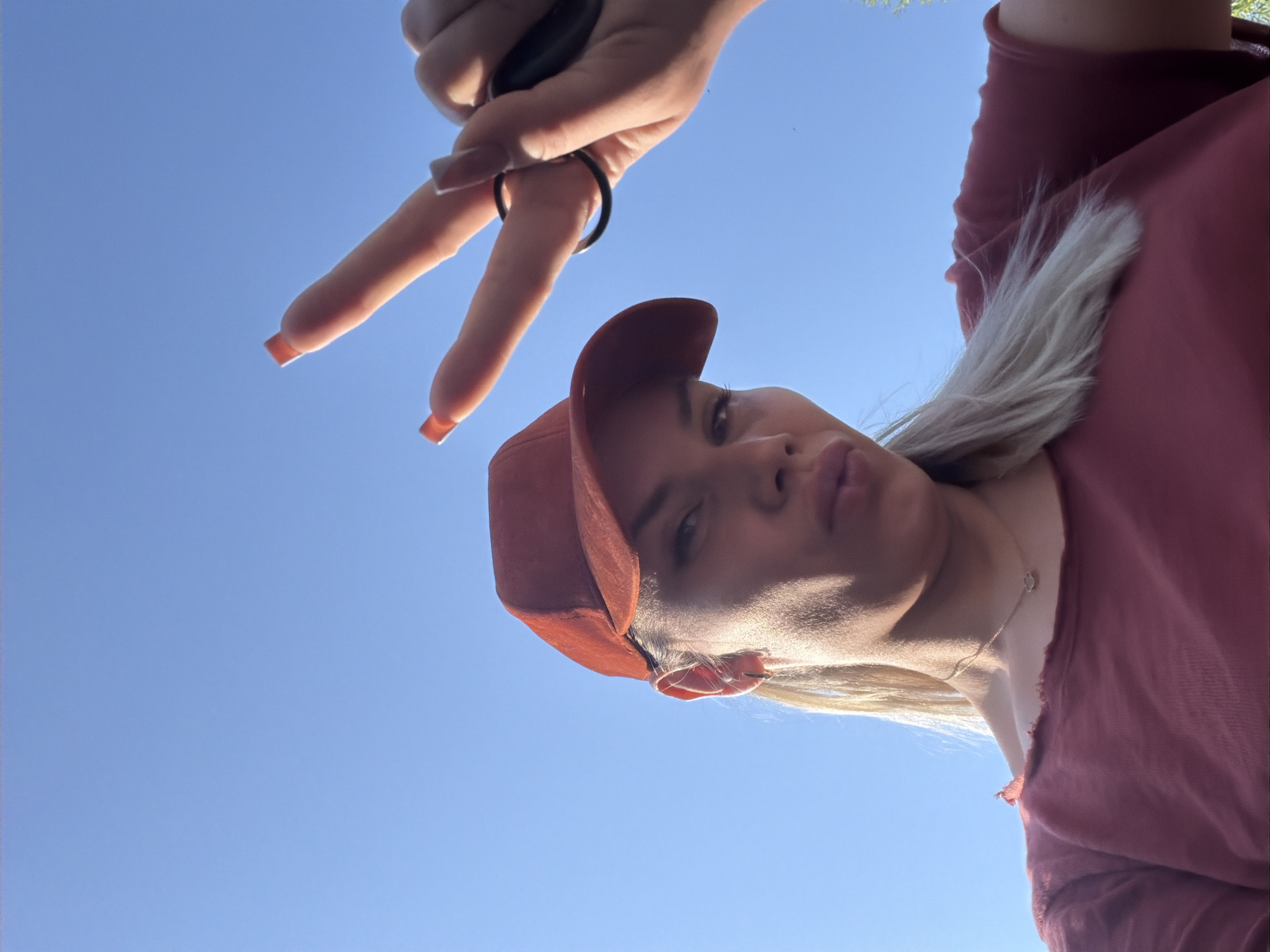 An outdoor, low-angle, close-up selfie shows a woman wearing a red baseball cap and a red t-shirt. She has blonde hair and is making a peace sign with her right hand, holding keys. The background is a clear blue sky. The image was taken in Moscow, Russia.