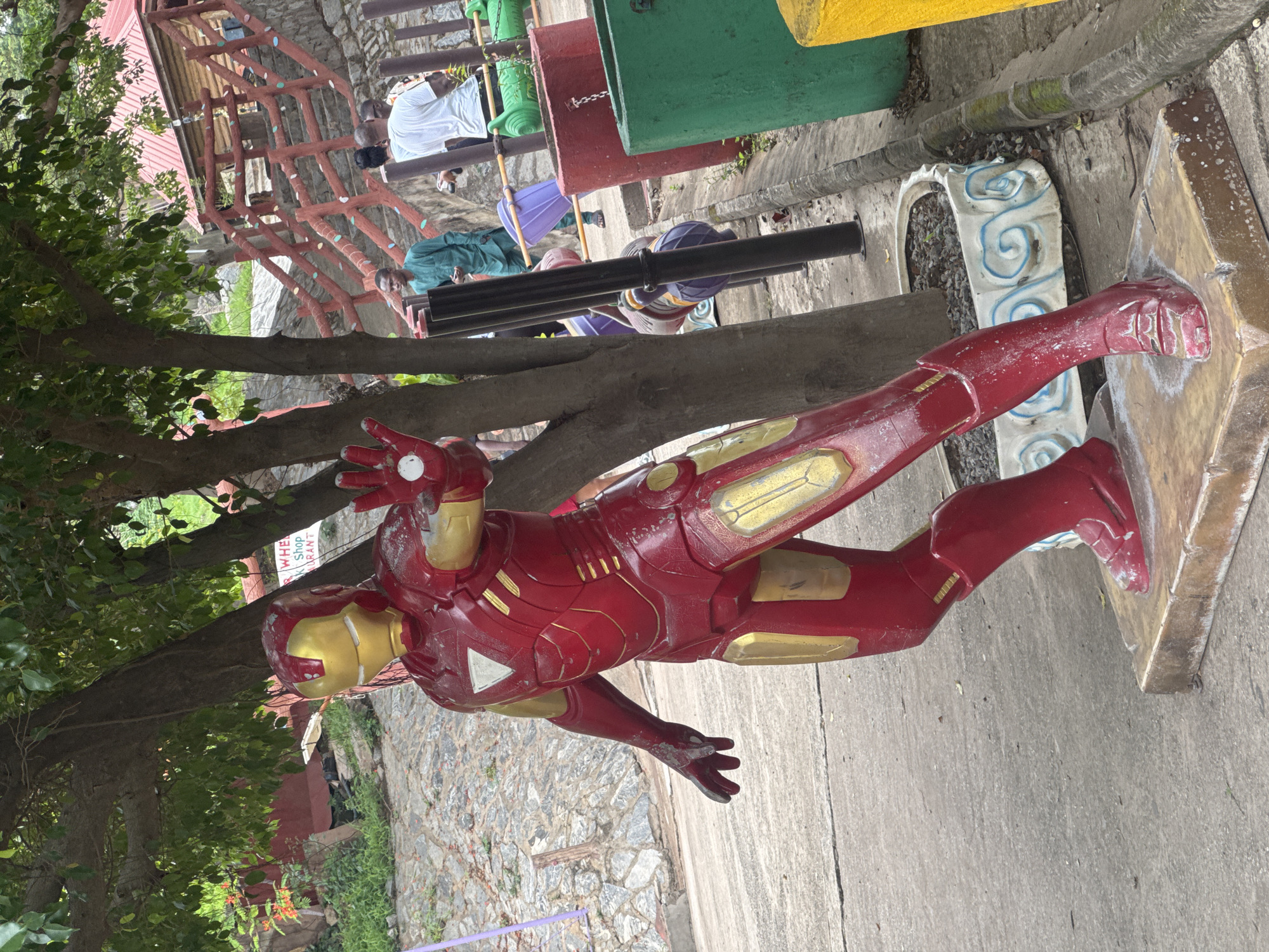 A life-size, slightly weathered, red and gold Iron Man statue stands in a public park in Abuja, Nigeria. The statue is posed mid-flight and mounted on a platform. Behind the statue is a playground structure, with individuals casually moving about, including one wearing a white shirt. A tree partially obscures the statue. The ground beneath the statue is paved.