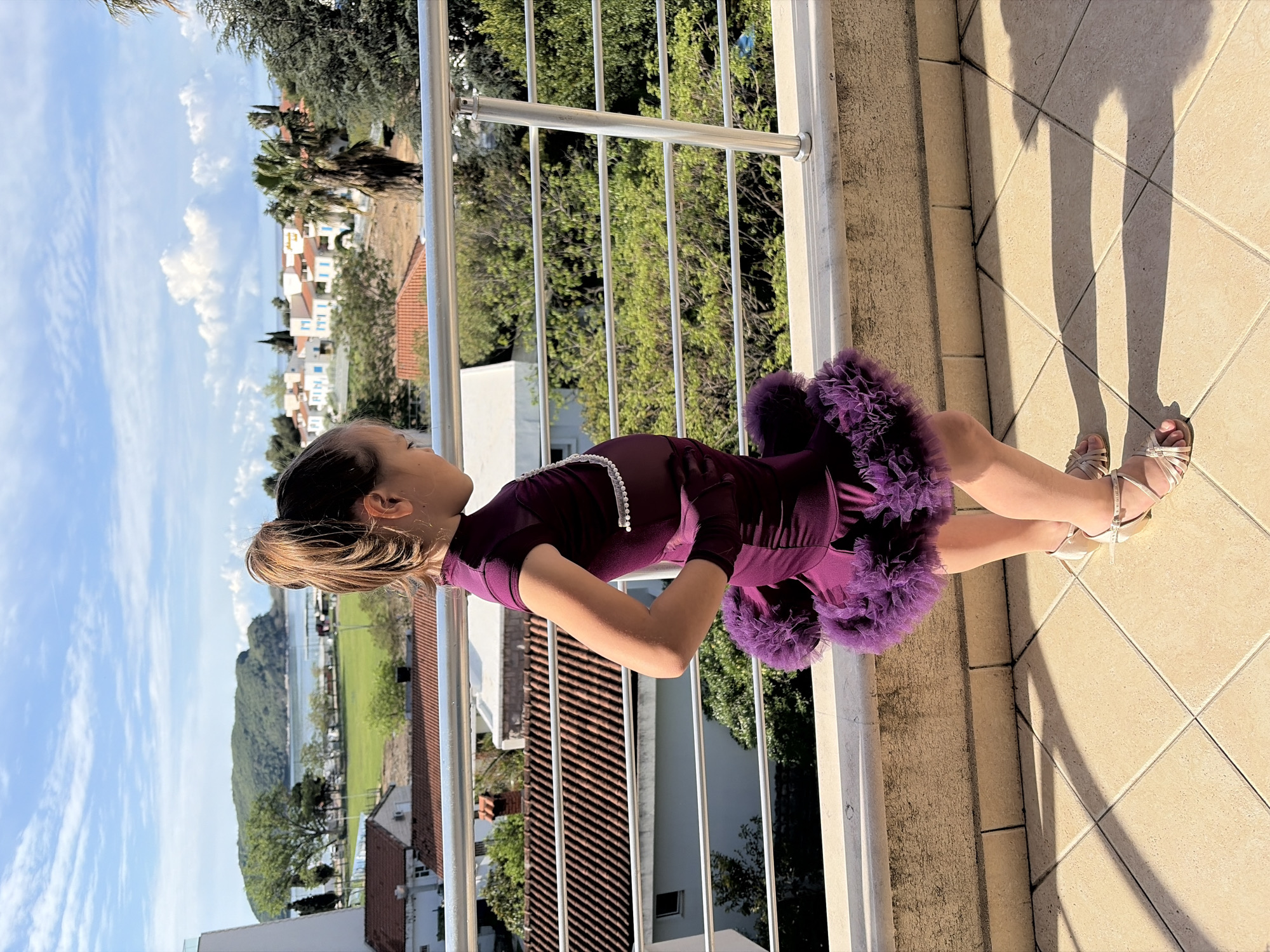 A young girl stands on a sunlit balcony, looking out at a scenic coastal town and natural landscape. She is wearing a shiny, dark purple long-sleeved dress featuring a voluminous, ruffled skirt of the same color. A white pearl-like necklace adorns her neck, and she also appears to wear matching dark purple gloves. Her light brown hair is tied back in a bun, and she has light-colored, strappy sandals on her feet.
The balcony floor is made of light-colored square tiles, and a silver metal railing separates the balcony from the view. Strong shadows, including those of her legs, are cast on the tiles, indicating bright, direct sunlight.
Beyond the railing, the background reveals a panoramic view characteristic of Budva, Montenegro. Numerous white buildings with distinct red-tiled roofs and some blue window frames are nestled among abundant green trees, including palm trees. In the distance, a body of water, likely a bay or the sea, stretches towards verdant mountains or hills. A green field is also visible closer to the water's edge. The sky above is clear blue with scattered white clouds. The scene suggests a bright, warm day, likely midday or early afternoon due to the angle of the shadows. No visible text is present in the image.