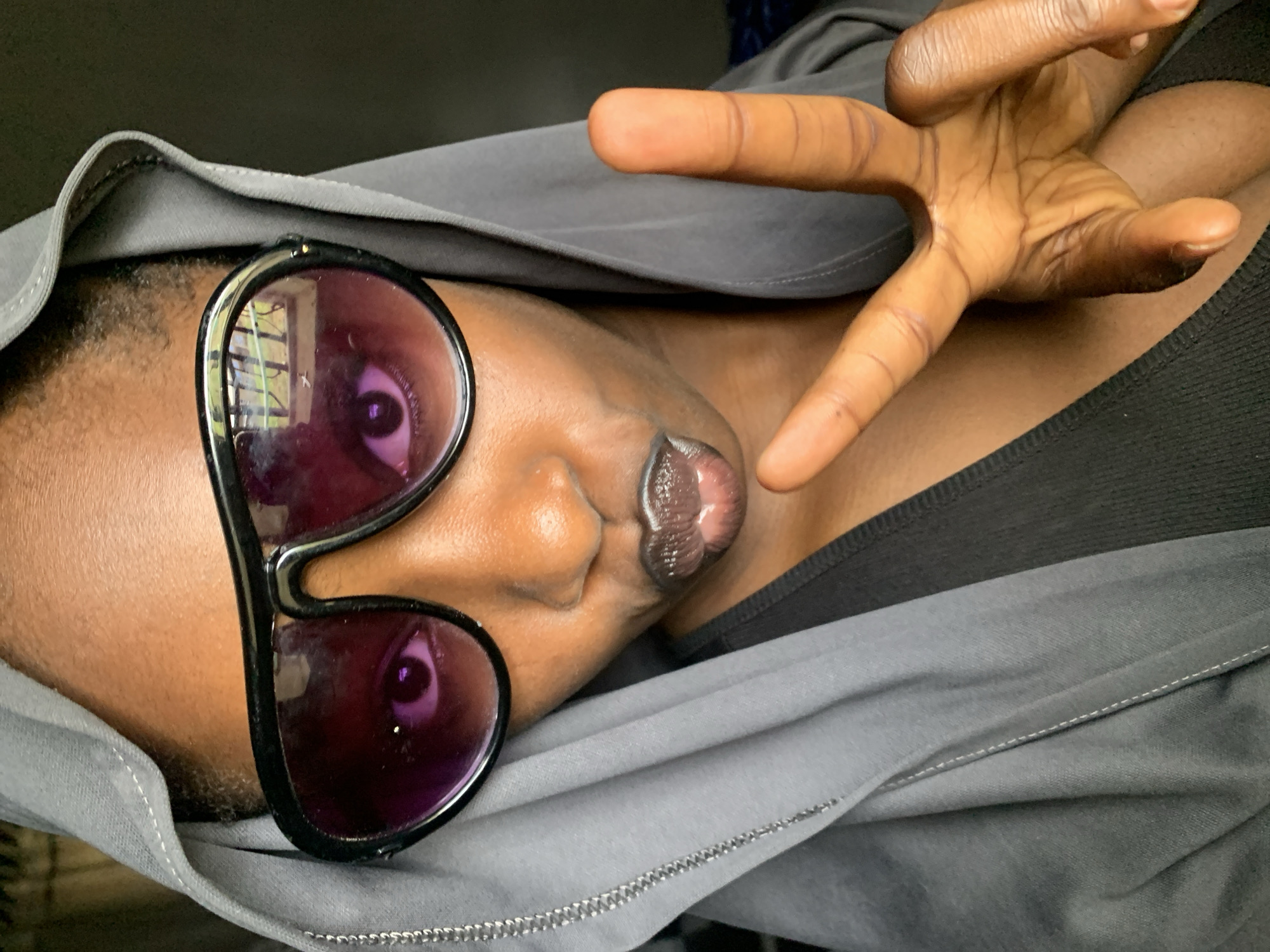 A close-up, overhead shot shows a Black person wearing a grey hooded garment and large, dark-rimmed sunglasses with purple lenses. Their lips are pursed, and they are holding up a peace sign with their right hand. The person's skin tone is brown. The background is dark. The image was taken in Rafin Par, Nigeria.
