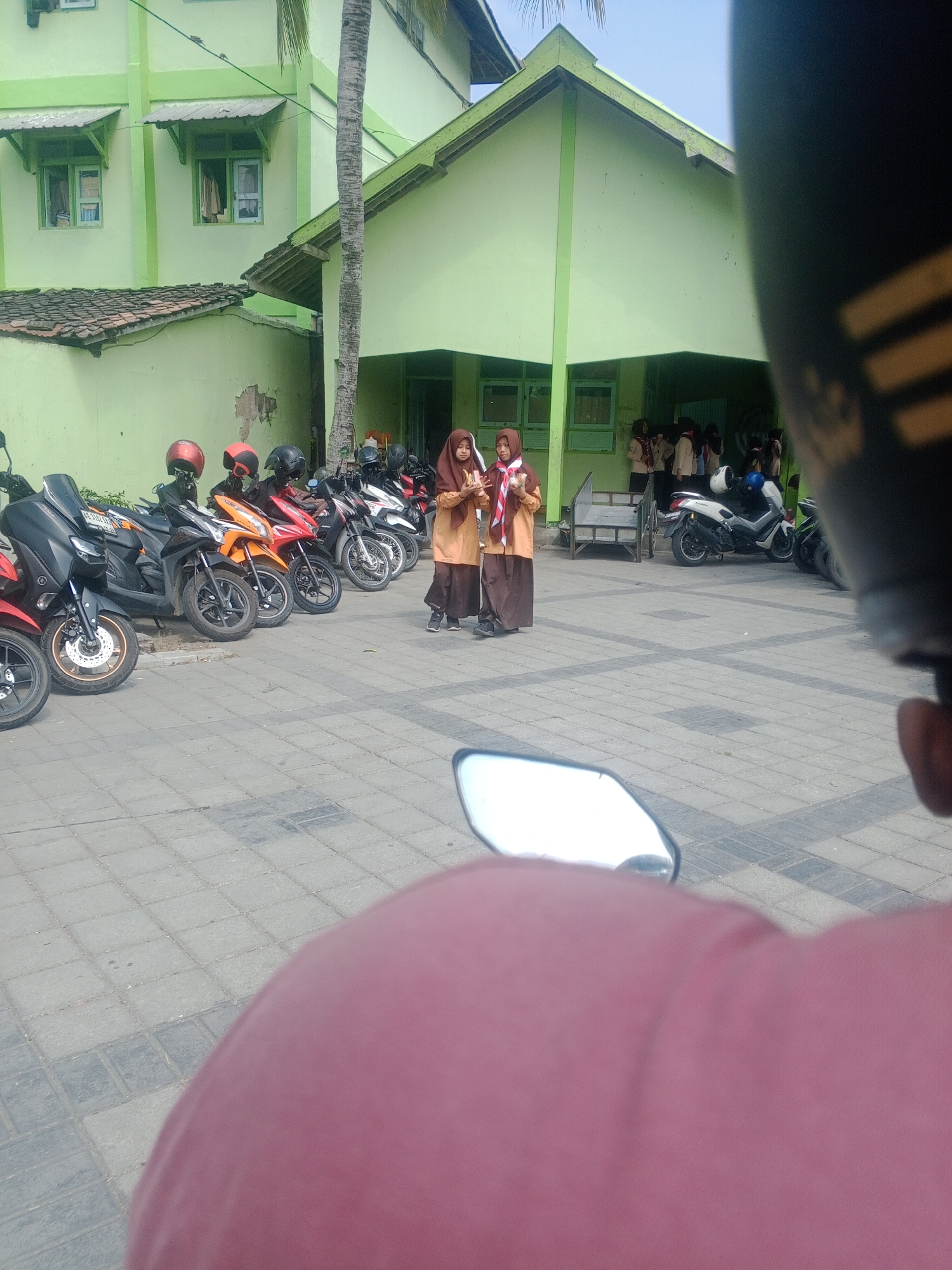 In Ronowijayan, Indonesia, a medium shot captures a group of uniformed students, identifiable by their hijabs and tan-colored uniforms, standing near a light green building. Two students in the foreground appear to be interacting, one holding a white and red banner or scarf. Behind them, a row of parked motorcycles, some with helmets on the handlebars, are visible in front of the building. The ground is paved with interlocking rectangular tiles. A palm tree trunk stands to the left of the students, and the building has a distinctive light green facade with a corrugated metal roof overhang. The image appears to be taken from the perspective of someone on a motorcycle, as a portion of a motorcycle helmet and a pink garment are in the foreground, obscuring part of the view.