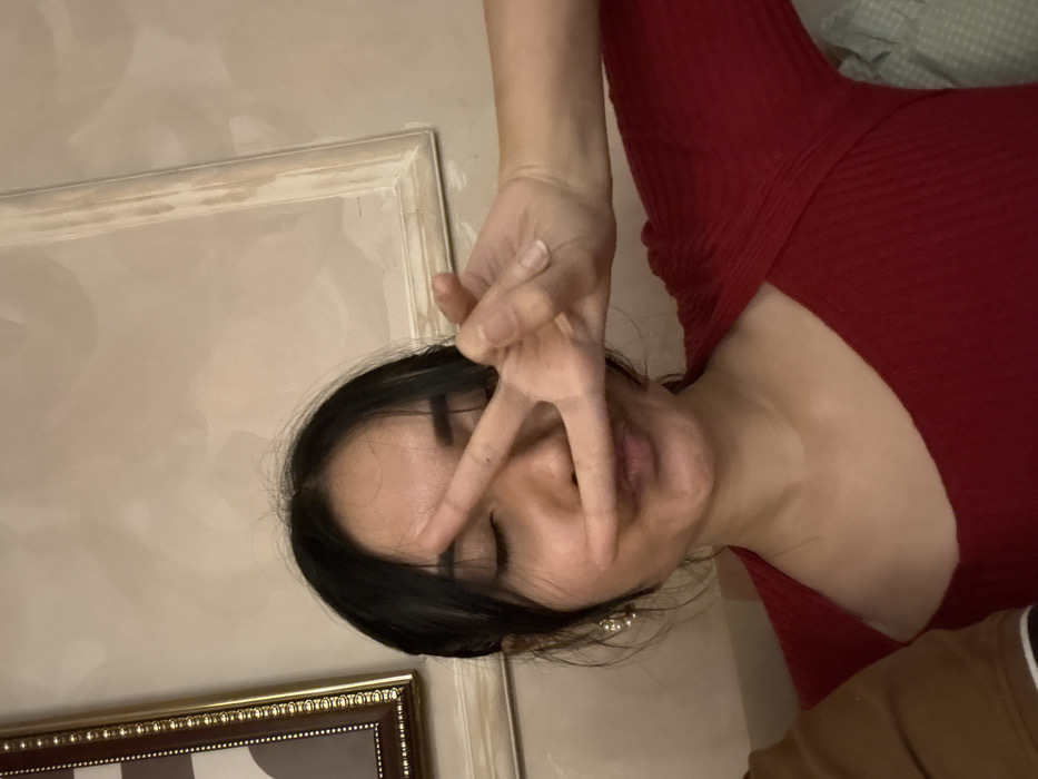 An Asian woman in a red dress poses with a peace sign partially covering her face. She has dark hair and her eyes are closed. Her complexion appears smooth, and a pearl earring is visible. The background features a light-colored wall, a decorative frame, and a glimpse of bedding, creating an indoor setting. The image is slightly tilted.