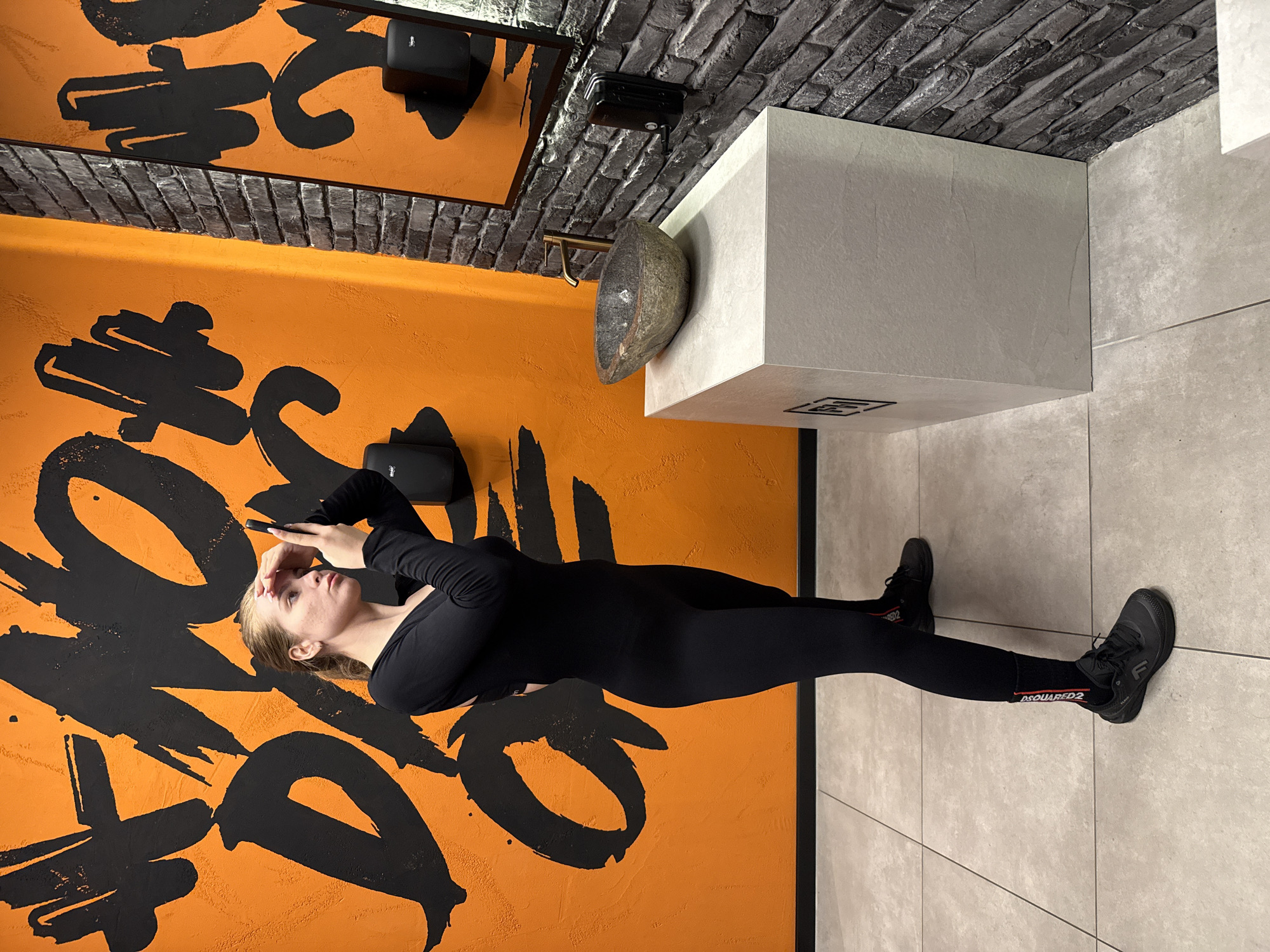 The image captures a woman standing in a modern indoor setting. She has blonde hair and is dressed in a black long-sleeved top, black leggings, black socks, and black sneakers. Her right hand is positioned near her forehead, while her left hand holds a dark, rectangular object, possibly a phone. She is looking slightly upwards and to her right.
The background features a striking, vibrant orange wall covered with large, abstract black graffiti-style brushstrokes. Above the orange wall, a dark, reflective surface, likely a mirror, shows reflections of the patterned wall. A section of dark gray brick-textured wall or ceiling is visible across the upper part of the frame. Two black rectangular devices, possibly speakers or network access points, are mounted on the wall or ceiling, one clearly showing a small white logo resembling a Wi-Fi symbol.
To the right of the woman, a light gray, cuboid structure, appearing to be a counter or vanity unit, is present. A unique, dark, irregularly shaped basin or decorative bowl is attached to its side, featuring a brass-colored faucet. The floor consists of light gray tiles.
Notable details include the red and white "DSQUARED2" text visible on the woman's black socks, and a stylized white 'F' logo on the side of her black sneakers. The scene presents a contemporary design with a strong visual contrast between the bold orange and black artistic wall and the sleek, muted gray architectural elements.