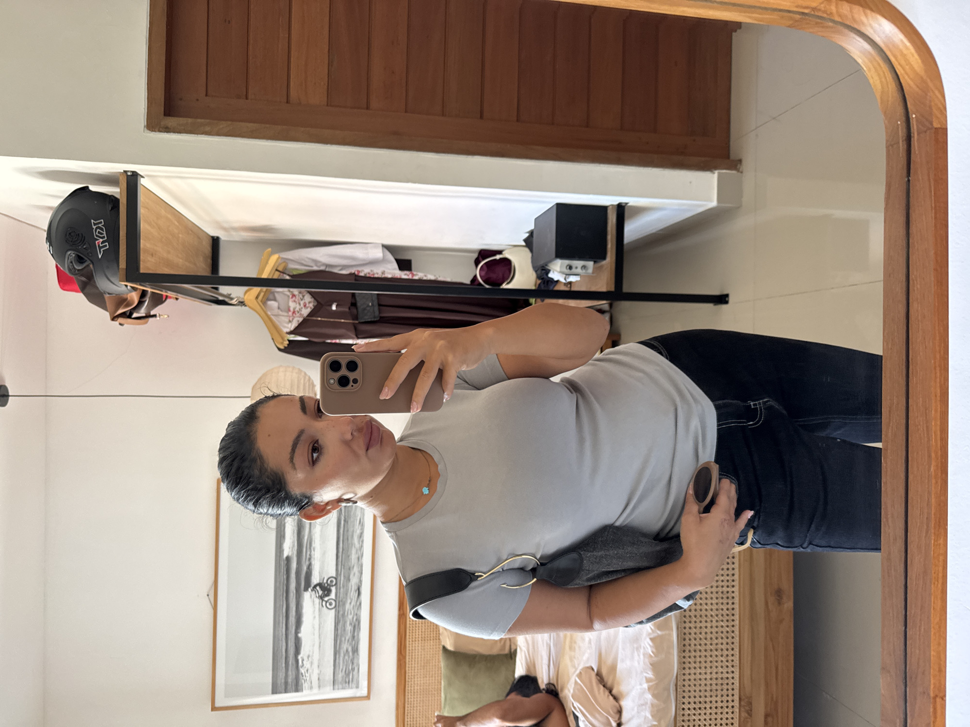This mirror selfie captures a woman indoors, rotated sideways. The subject is a woman with dark hair pulled back, wearing a light grey t-shirt and dark jeans. She holds a light-colored smartphone in her right hand to take the photo, looking towards her reflection with a neutral expression. A small, light blue clover-shaped pendant is visible around her neck, and she wears a dark grey or black shoulder bag with a prominent gold circular buckle. Her left hand rests on her hip, holding what appears to be a pair of sunglasses.
The reflection reveals details of the room. On the left, behind the woman, a wooden bed frame with a woven rattan headboard is partially visible, along with bedding. Above the bed, a framed black and white photograph depicts a motorcycle on a beach. On the right, a black metal and wood shelving unit holds a black motorcycle helmet with "KYT" written in red and white on its side. Below the helmet, various clothes hang on the rack, and a black box or device is on a shelf. The room features white walls, wooden slatted wall panels, and what appears to be a wooden door or cabinet. The mirror itself has a thick, curved wooden frame. The lighting suggests it is daytime. The interior design elements, including the rattan and wood, are consistent with a location in Kuta Selatan, Indonesia.