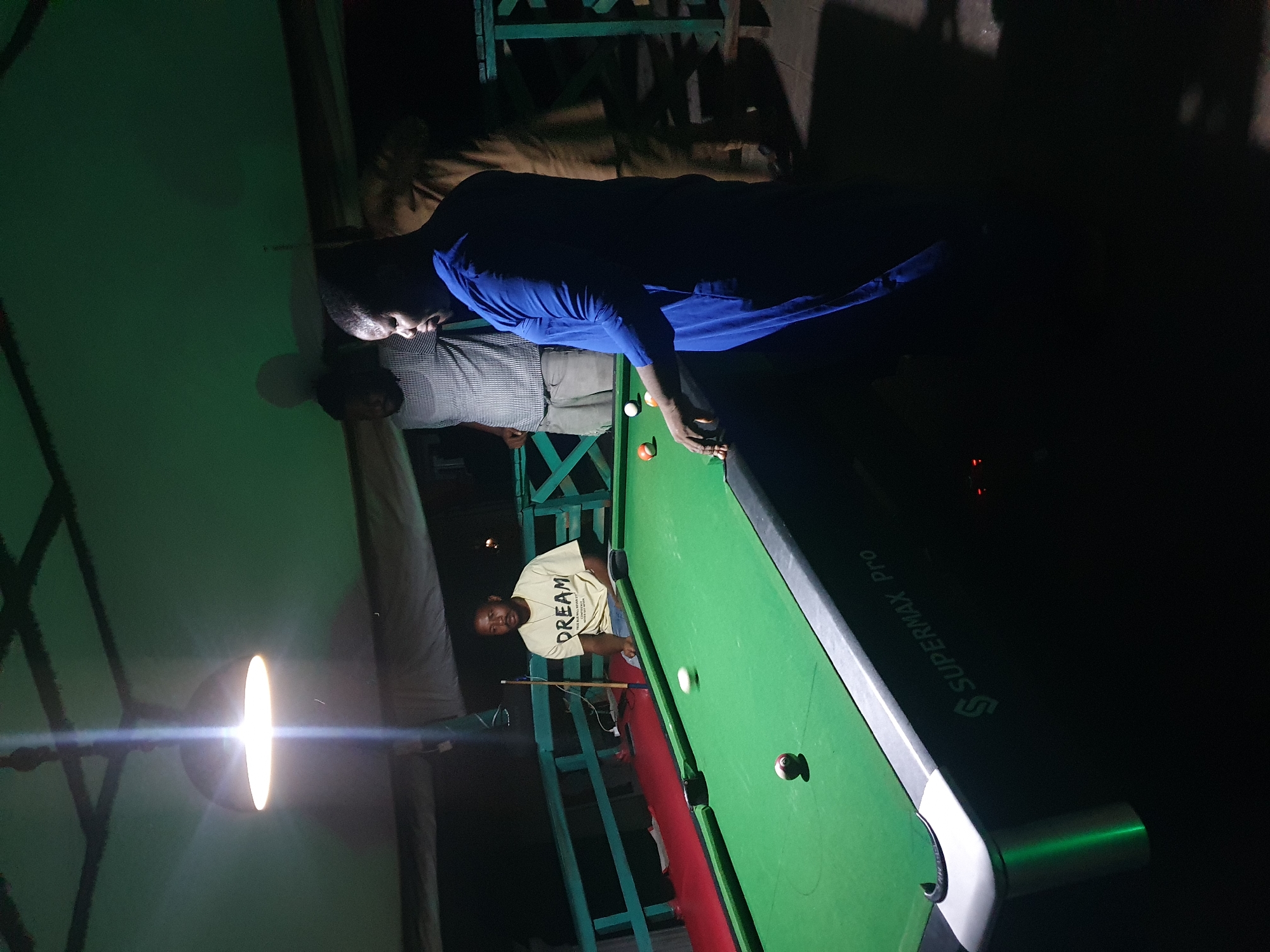 In Lokoja, Nigeria, a dimly lit room with a green ceiling features a group of men gathered around a pool table. A bright overhead light illuminates the scene. Three men are visible playing pool. One man in a blue outfit stands on the right, facing away from the camera, engaged in the game. Another man, wearing a light-colored, striped shirt, stands behind him. A third man, in a yellow t-shirt with the word "DREAM" printed on it, is seated on the left side of the pool table. Several pool balls are scattered on the green felt surface, with one white ball in motion. The pool table is branded "SUPERMAX Pro." The environment appears to be an indoor recreational space, possibly a bar or club, with a railing visible in the background.