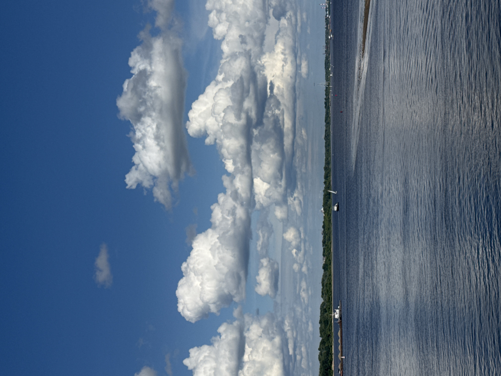 This is a vertical, medium-shot photograph of a wide river under a bright blue sky with scattered white clouds. The foreground is dominated by the dark blue water, with subtle ripples on the surface. In the middle ground, a long, brown barge carrying what appears to be sand or gravel is traveling from left to right, accompanied by a smaller white boat. Further to the right, a white sailboat is visible on the water. The background features a lush green shoreline with dense trees, suggesting a park or forested area. Beyond the treeline, distant industrial structures and buildings are faintly visible. The scene is devoid of any people. The perspective suggests the photo was taken from another boat or from a high vantage point near the river. The overall atmosphere is one of peaceful activity on a clear day. This scene is likely the Volga River in Nizhny Novgorod, Russia, given the presence of barges, industrial elements, and the characteristic wide river expanse.