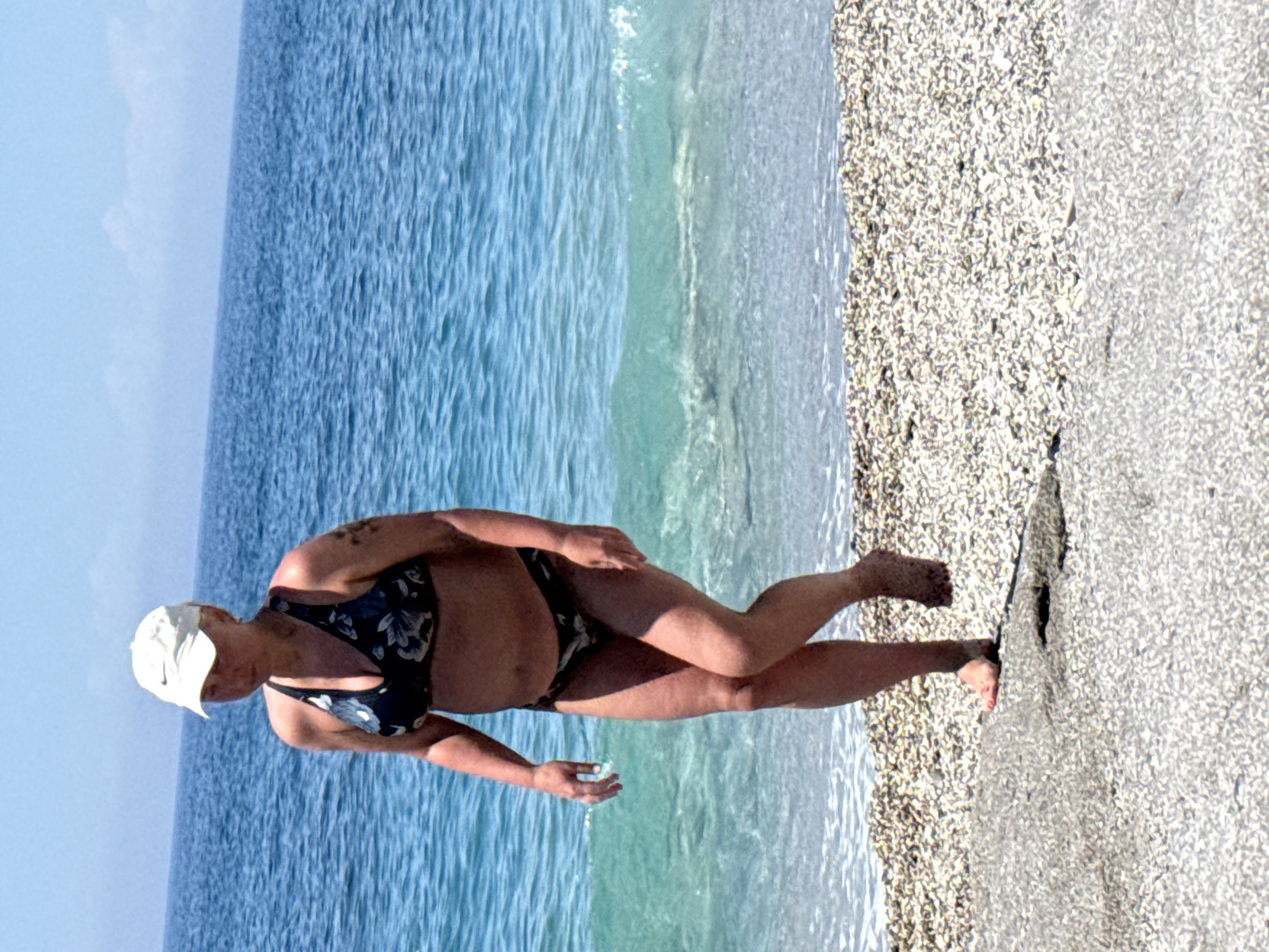 A woman in a floral bikini and white cap stands on a beach with one foot in the water and the other on the sand. The water is blue and clear, and the sky is light blue. The beach is made of small pebbles. The location is Unknown, Unknown.
