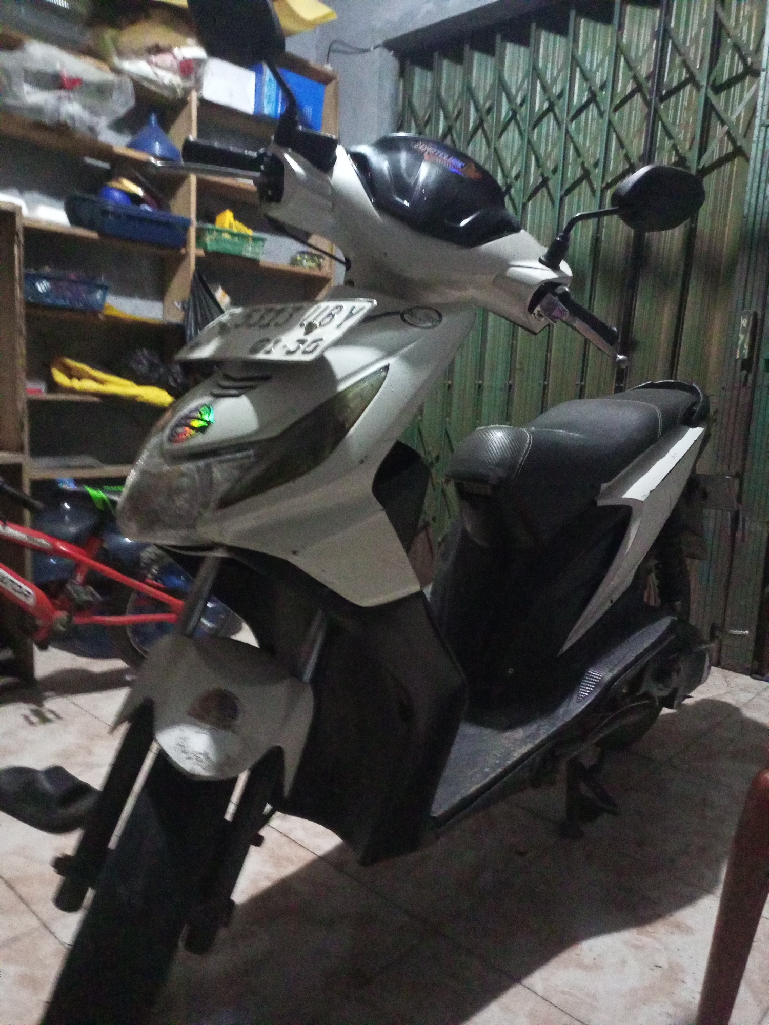 The image shows a white scooter parked indoors, likely in a garage or storage area, with shelves filled with various items in the background. A collapsible green metal gate is visible behind the scooter. The scooter has a license plate with the text "B 3333 UBY" and "01-35". A red bicycle is partially visible to the left of the scooter. The flooring is tiled. The scene suggests a residential or storage setting in Cidahu, Indonesia.