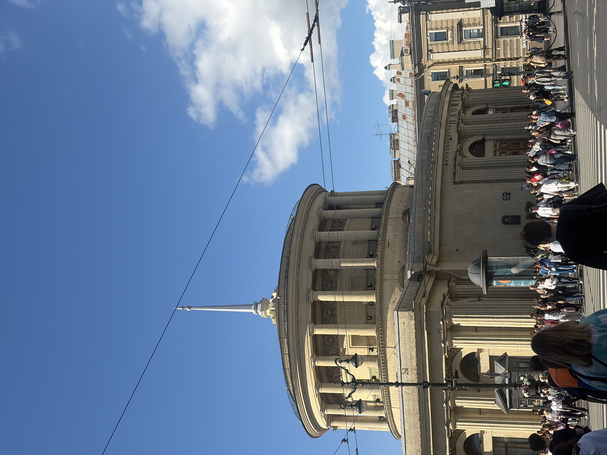 A low-angle shot captures the facade of the Saint Petersburg Metro station, "Ploshchad Vosstaniya," under a bright blue sky with scattered clouds. The building is a neoclassical rotunda with a colonnade of tall, fluted columns supporting a circular entablature and balustrade. A slender, pointed spire rises from the center of the roof. In front of the building, a black ornate street lamp stands prominently. A busy crosswalk is filled with pedestrians of diverse ages, dressed in casual summer attire, crossing the street. To the right of the Metro station, a multi-story building with numerous windows and an ornate facade is visible, along with rooftop structures. Overhead, power lines stretch across the sky, and an overhead traffic signal displaying a green light is visible. The overall impression is a bustling urban scene on a clear day.