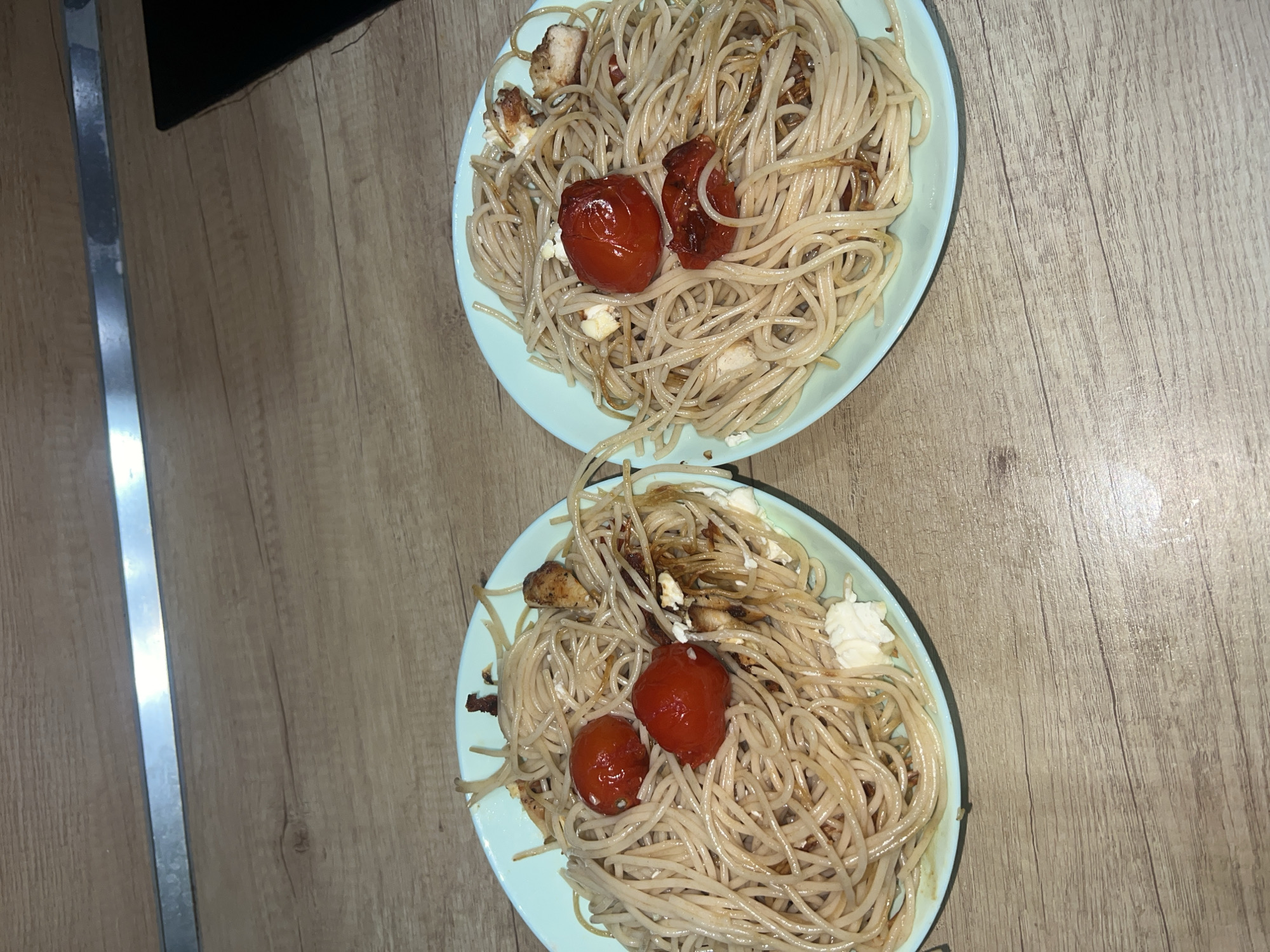 Two plates of spaghetti with tomatoes and chicken are on a wood-patterned surface. The location is not discernible from the image.