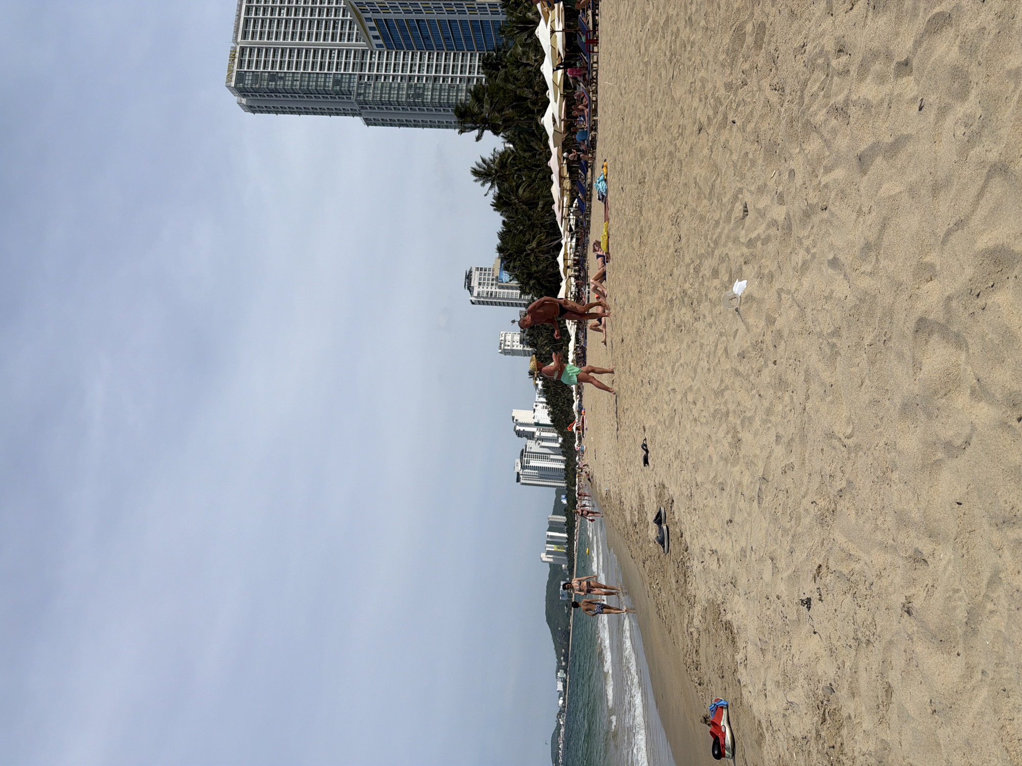 A wide, sandy beach is pictured in Nha Trang Ward, Vietnam. The ocean is on the left, with gentle waves. In the background, several high-rise buildings line the coast. People are visible on the beach, some walking, others relaxing on lounge chairs under umbrellas. The sand is marked with footprints and scattered with a few items of clothing and footwear. A piece of white trash is visible on the sand in the mid-ground. The sky is overcast.