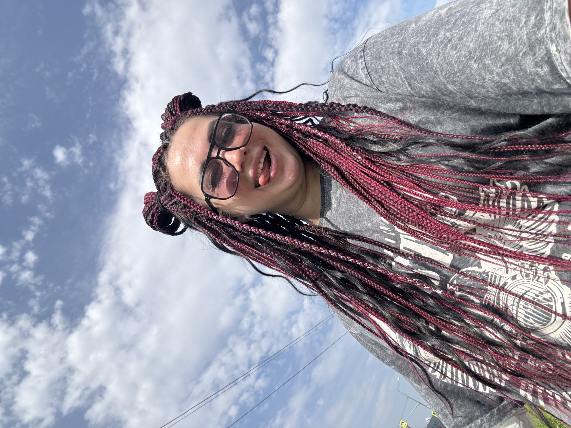 A young woman with dark skin, dark eyebrows, and dark hair styled in long braids with burgundy highlights, and two top knots is shown from the chest up, taken from a low angle. She is wearing large, black-rimmed eyeglasses, and a grey t-shirt with a graphic design. She is sticking her tongue out and smiling. The background is a bright blue sky with white clouds and some telephone wires. The image was taken outdoors in Tolyatti, Russia.
