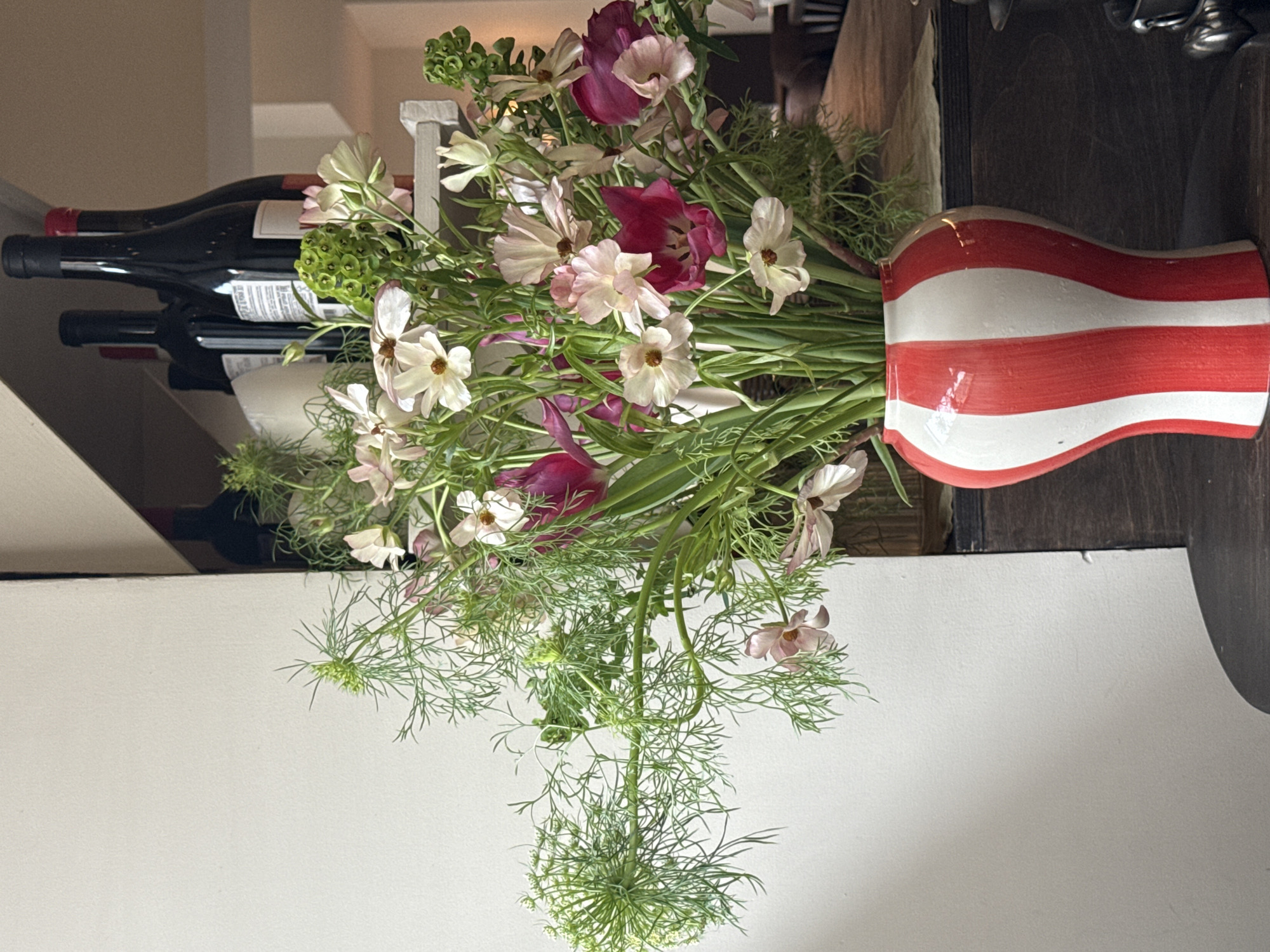 A vase of flowers sits on a dark surface, likely a countertop or table. The vase is red and white striped and bulbous. It contains a variety of flowers, including white and pink ones, as well as greenery. Behind the vase, blurred in the background, are wine bottles on a rack, a wooden board, and some utensils. The lower part of the image has a white wall or surface. The scene appears to be indoors, possibly in a kitchen or dining area. There is no evidence to suggest that the scene is in Moscow, Russia.