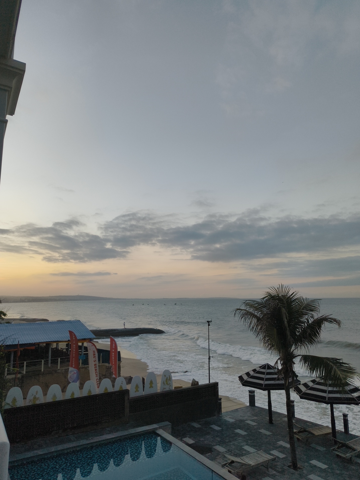 This media file depicts a tranquil scene at Phu Thuy Ward, Vietnam, during what appears to be sunrise or sunset. The sky is a soft gradient of grey and pale yellow, with scattered clouds. The main subjects are the ocean and the beach, with gentle waves rolling onto the shore.
In the foreground, a swimming pool with a tiled bottom is visible, reflecting the sky. To the right, a paved patio area features two striped beach umbrellas and lounge chairs, suggesting a resort or hotel setting. A palm tree stands tall, its fronds reaching towards the sky.
Further back, a beachside establishment with a blue roof is discernible, with several flags displaying text. One flag reads "JIBE'S BEACH CLUB". There are also multiple surfboards lined up, indicating a potential surf school or rental. A few small boats are scattered on the water in the distance.
The overall mood is peaceful and serene, capturing the quiet beauty of a coastal location. The time of day is suggested by the soft lighting and the calm atmosphere. There are no prominent human figures, except for a solitary person on a rock formation by the water. The weather appears to be clear and calm.