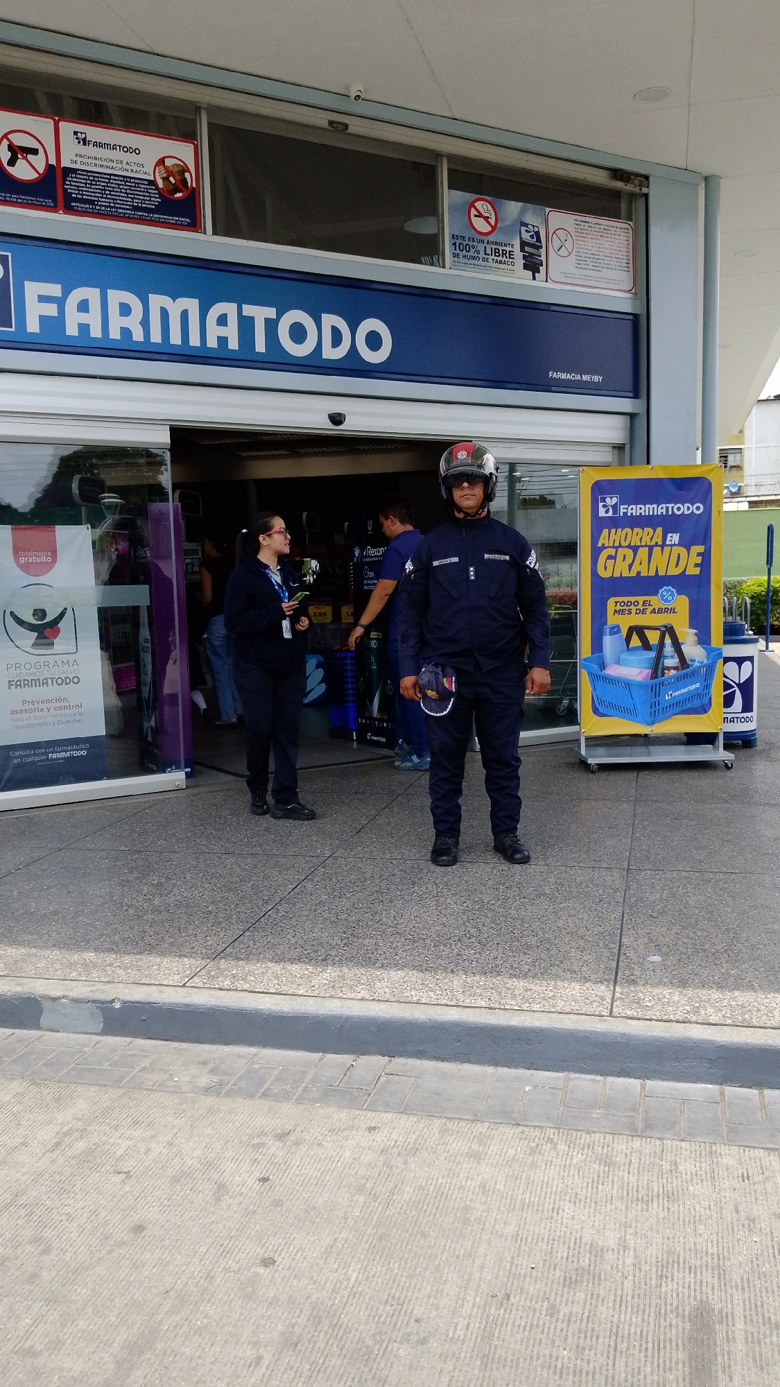 The image captures the entrance of a "FARMATODO" pharmacy in Tinaquillo, Venezuela, during daytime. A security guard, a man wearing a dark blue uniform, black boots, dark sunglasses, and a motorcycle helmet, stands prominently in front of the automatic glass doors. A blue and white baseball cap is tucked into his front pocket.
To his left, a female employee in a dark blue uniform (shirt and pants) and glasses is visible near the entrance, looking towards the inside of the store, possibly assisting customers or working. Other customers are partially visible inside the pharmacy.
The pharmacy's name, "FARMATODO," is displayed in large blue letters above the entrance. Several informational signs are affixed to the glass facade above the doors. These include a "PROHIBICIÓN DE ACTOS DE DISCRIMINACIÓN RACIAL" (Prohibition of Acts of Racial Discrimination) sign on the left, and an "ESTE ES UN AMBIENTE 100% LIBRE DE HUMO DE TABACO" (This is a 100% Tobacco Smoke-Free Environment) sign on the right. A smaller sign above the right door indicates "FARMACIA MEYBY."
To the left of the entrance, a vertical banner promotes the "PROGRAMA CUIDAMOS TU SALUD FARMATODO," offering "Prevención, asesoría y control para el tratamiento de la Hipertensión y Diabetes," and advising to "Consulta con un farmacéutico en cualquier FARMATODO." On the right, a tall yellow vertical sign with "FARMATODO" branding advertises "AHORRA EN GRANDE" (Save Big) "TODO EL MES DE ABRIL" (All of April), featuring a graphic of a blue shopping basket filled with products. The overall setting appears bright and clear under sunny weather.