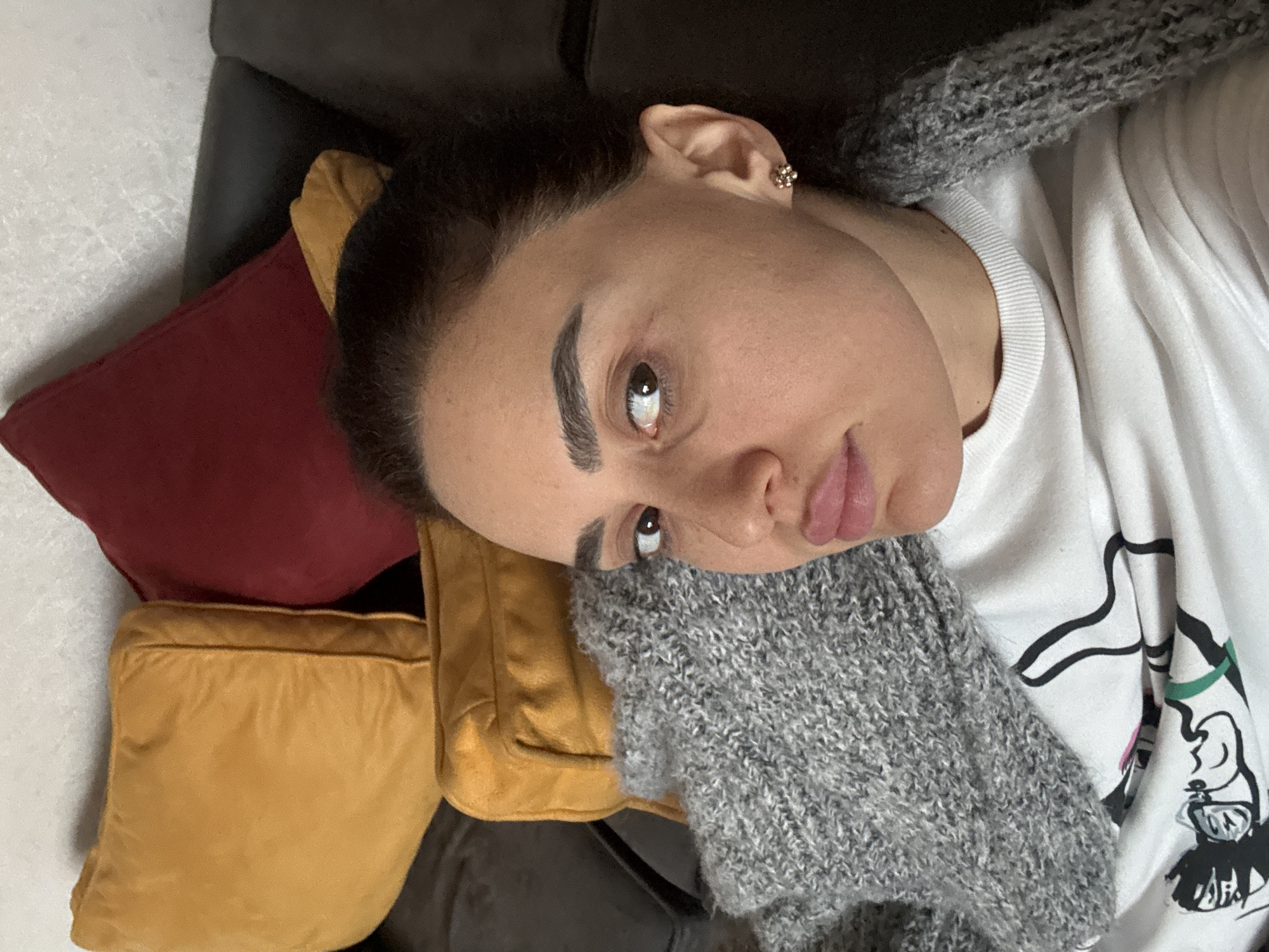 A woman is lying on a dark-colored sofa with several pillows. There are two yellow pillows and one red pillow near her head. She is wearing a white shirt with a black design and a grey knitted garment, likely a scarf or sweater, around her neck. She has dark hair pulled back, visible eyebrows, and wears an earring. The location is not discernible.