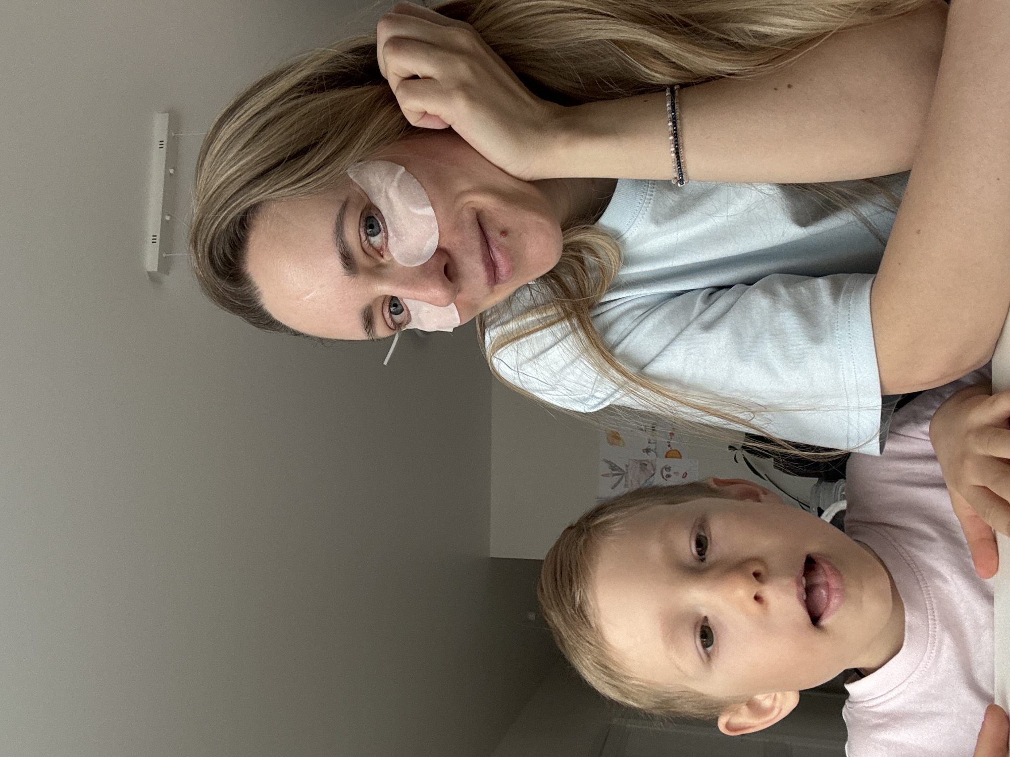 A woman and a young child are visible in this image. The woman, with blonde hair, is wearing eye masks. She also has a bracelet on her wrist. The child has light blonde hair and appears to be sticking out their tongue. They are both wearing t-shirts. The setting appears to be indoors, possibly a home in the city of Chaikovsky, Russia. Details on the wall behind them are not readily identifiable.