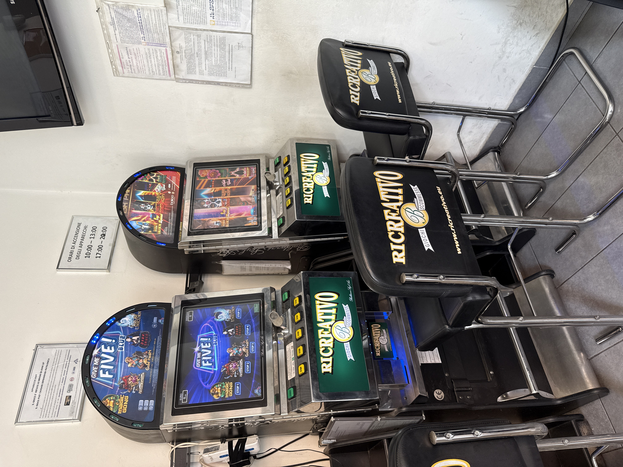 The image shows two slot machines, branded "GIVE ME FIVE!", with a gaming chair in front of each. The machines are silver and black, with a colorful display screen showing game graphics. The chairs are black leatherette with chrome legs and feature the "RICCREATIVO" logo. In the background, there are several posters on the wall, including one with opening hours for the machines and another detailing rules and regulations for gambling. The environment appears to be an indoor gaming or entertainment establishment. The floor is tiled with grey tiles. Based on the Italian language used on the signs and the branding, it is likely a location in Italy. Without more specific visual cues like street signs or unique landmarks, it's impossible to pinpoint the exact location within Bologna, Italy, although the overall context suggests a gaming establishment within that city.