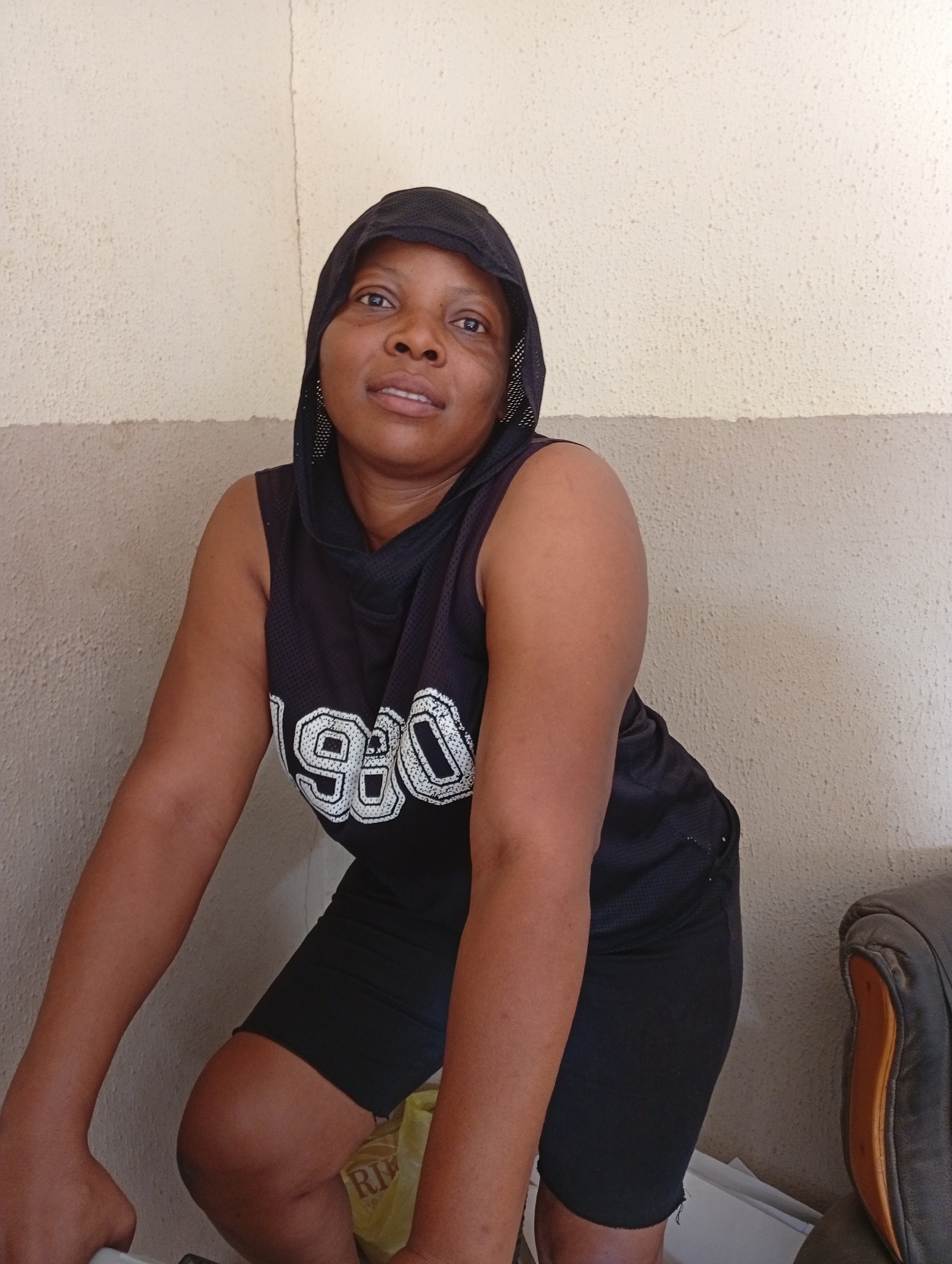 A medium shot depicts a dark-skinned woman wearing a black hooded tank top with the number "980" printed in white on the front, and black shorts. She is indoors, positioned in front of a two-toned wall with a tan upper section and a grey lower section. A chair with a brown armrest is partially visible on the right side of the frame. Based on the information provided, the location is Sabon Tasha, Nigeria, but no features specific to that city are present. The woman is looking at the camera. No further actions or interactions are observable. The environment consists of the immediate interior space with its painted wall.