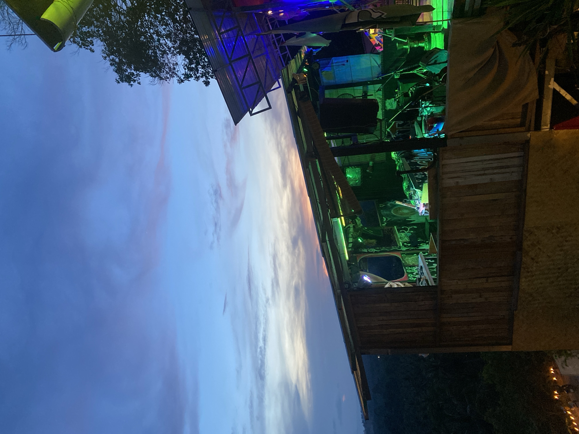 The image shows an outdoor scene in Baan Don Sai, Thailand. A structure, possibly a bar or stage, is partially visible on the right side of the frame, constructed of wood and illuminated with green and blue lights. The sky occupies the left portion of the image, displaying a gradient of blue and pink hues at dusk or dawn. Trees are visible in the background. The image is oriented vertically.