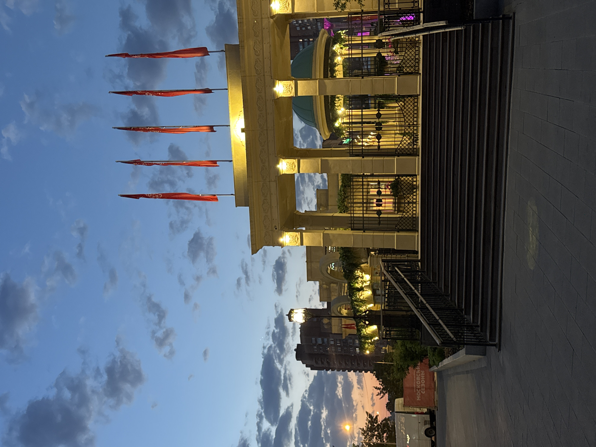 The image shows a building with red flags and architectural details in Moscow, Russia. The building features ornate columns, yellow lighting, and a green dome visible through the arches. There are a series of red flags hanging vertically from poles on the left side. The sky is blue with grey and white clouds, suggesting it is either dusk or dawn. There is a set of stairs leading up to the building, with metal railings on the sides. The ground is paved with dark grey tiles. In the background, another building is partially visible, along with trees and a street light. The red text visible on the lower right side reads "Московский сезоны" (Moscow Seasons), which is further confirmed by the location of Moscow.