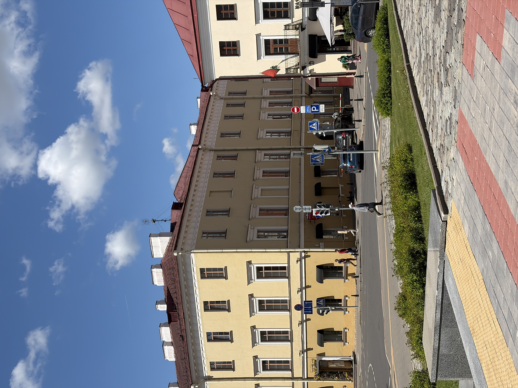 The image depicts a street in Minsk, Belarus. The buildings are multi-storied, painted yellow, and have red roofs. Signage is in evidence. A street is in the middle of the image. Pedestrians are visible. The sky is blue with white clouds. The sidewalk is paved with red and yellow blocks. A small patch of greenery separates the street and sidewalk. Cars are parked along the street.