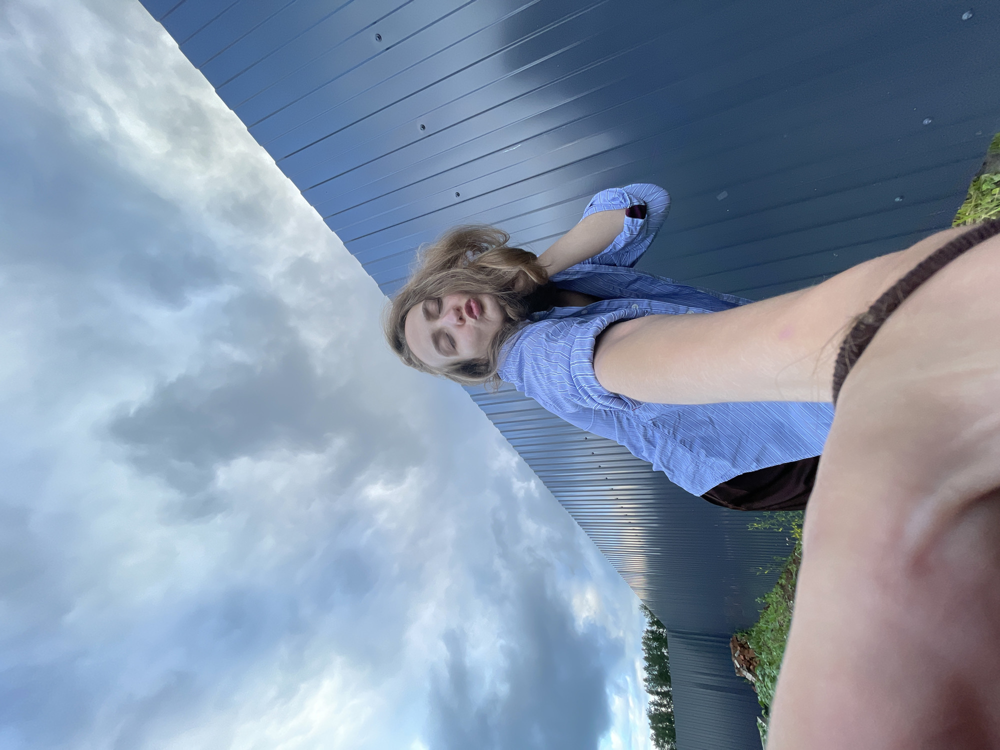 A young woman with blonde hair is taking a selfie outdoors in a rural setting, possibly in Podshivalovo, Russia. She is wearing a blue striped button-down shirt and dark shorts. Her eyes are closed, and her lips are puckered as if she is blowing a kiss. She is leaning against a tall, dark blue corrugated metal fence. The sky above is filled with dramatic, grey and white clouds. In the background, a line of trees is visible on the left side of the image. The ground in front of the fence is covered in green grass and some scattered debris, including a small pile of bricks. The woman's arm is extended, holding the camera.