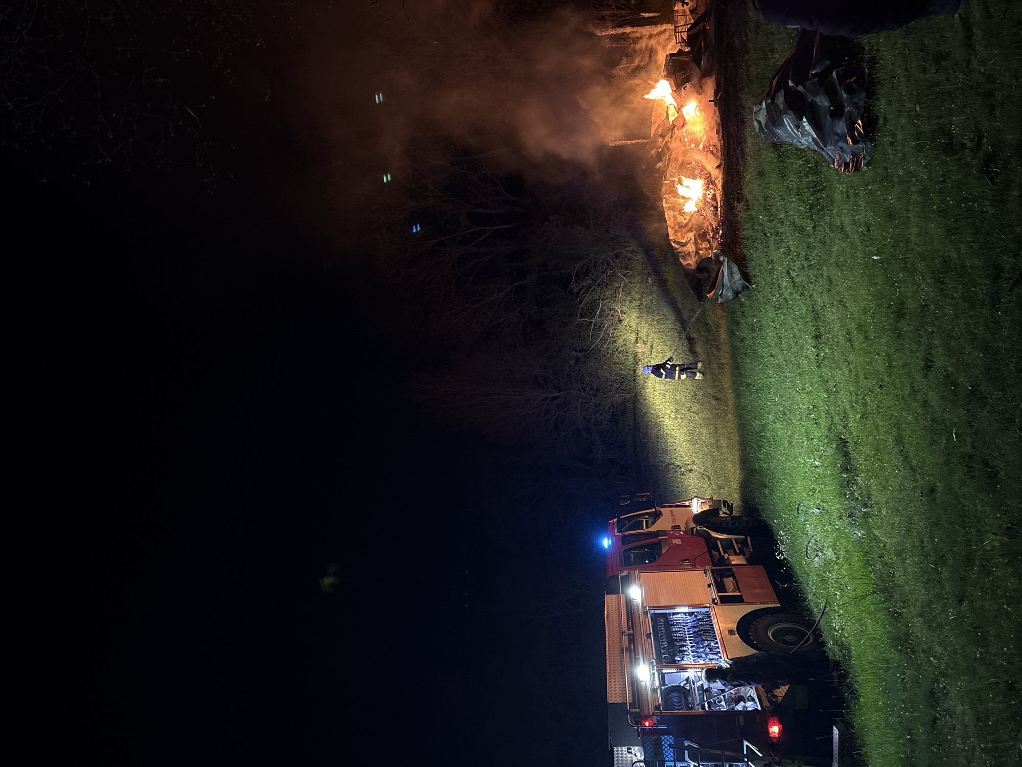 The image captures a nighttime scene in Dolno Drianovo, Bulgaria, dominated by a burning fire and the presence of emergency services.
In the foreground, a large fire is ablaze, emitting bright orange and yellow flames and a significant amount of smoke, which glows orange from the firelight. The fire appears to be consuming a pile of debris or other material, leaving charred remnants. Near the fire, a firefighter stands, illuminated by the flames and other light sources. This individual is wearing a protective suit, blue on the top half and yellow on the bottom.
To the right of the scene, a red and white fire truck is parked on grassy ground. Its emergency blue lights are active, casting a blue glow on the immediate surroundings. The truck's compartments are open, revealing various pieces of equipment and hoses. A hose can be seen extending from the truck towards the area of the fire, indicating active fire suppression or monitoring. Another person, likely a firefighter, is visible near the back of the truck. The side of the fire truck features partial text, including "ПОЖАРНА" (Fire Department) and possibly "ДЕЖУРЕН".
The entire scene is set outdoors at night. The ground is covered in grass, and bare tree branches are visible against the dark sky in the background, partially obscured by smoke. The only significant illumination comes from the intense flames of the fire and the spotlights emanating from the fire truck, casting harsh shadows and highlighting areas of interest. The overall impression is one of an ongoing emergency response to a fire incident.