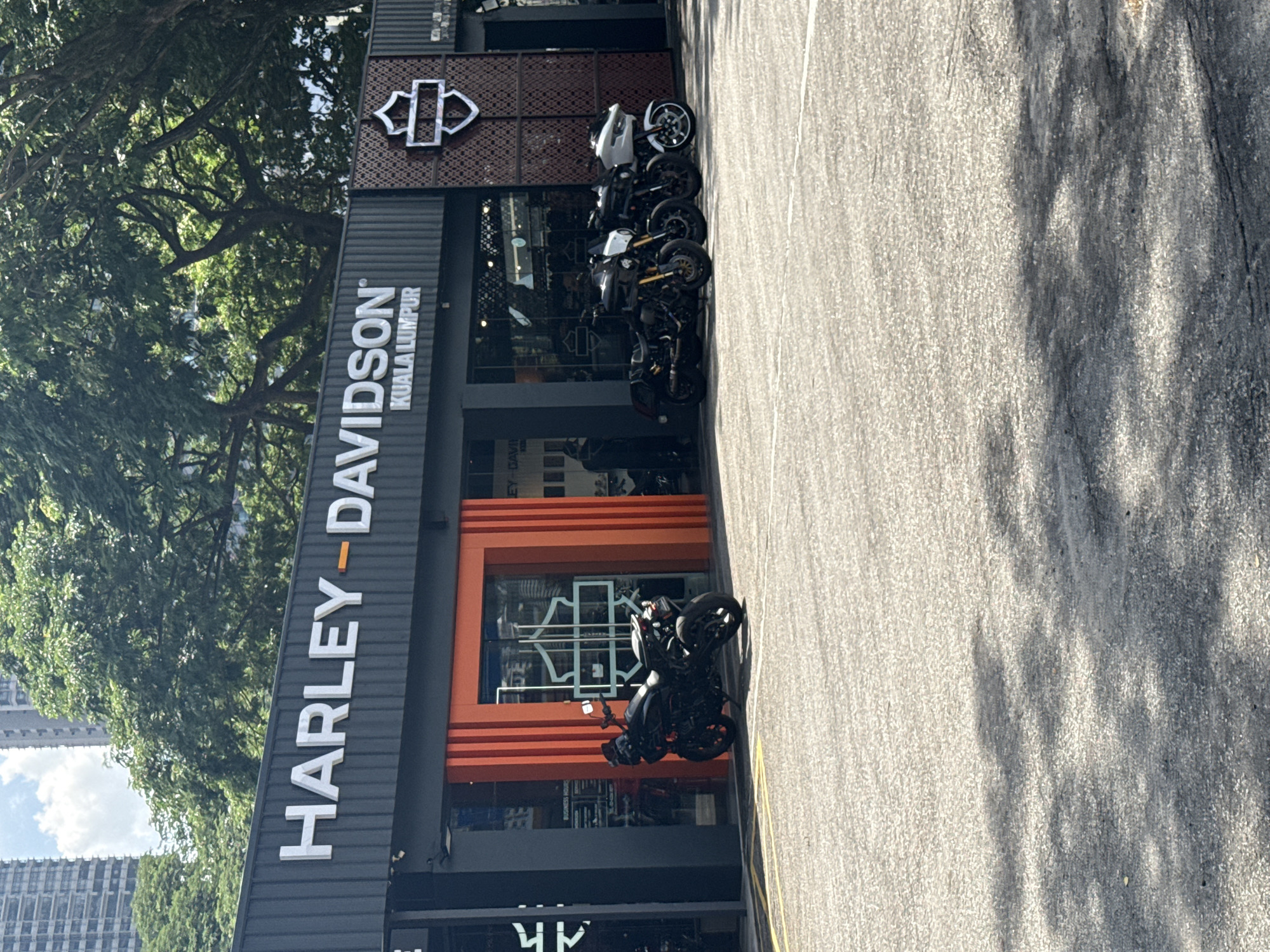 The image displays an outdoor scene of a Harley-Davidson motorcycle dealership in Kuala Lumpur, Malaysia, under bright, sunny conditions. The main subject is a modern, dark-grey building featuring large, white illuminated lettering that spells "HARLEY-DAVIDSON KUALA LUMPUR" on its facade. The distinctive Harley-Davidson bar and shield logo is prominently visible above the main sign and on a decorative, textured panel to the right.
In front of the dealership, a light grey paved area, likely an asphalt parking lot, stretches horizontally. Several motorcycles are parked on this surface near the building; at least four are clearly visible, primarily in dark colors with some silver or white accents. The dealership's main entrance is highlighted by bold orange horizontal panels surrounding large glass windows, through which interior branding is faintly visible.
Lush green trees with dense foliage are abundant to the left and behind the dealership building, suggesting a vibrant urban green space. In the background, glimpses of tall, contemporary city buildings can be seen under a partly cloudy blue sky. The strong sunlight casts distinct shadows on the paved ground, indicating a midday or early afternoon setting. The scene is static, focusing on the commercial establishment and its display of motorcycles.