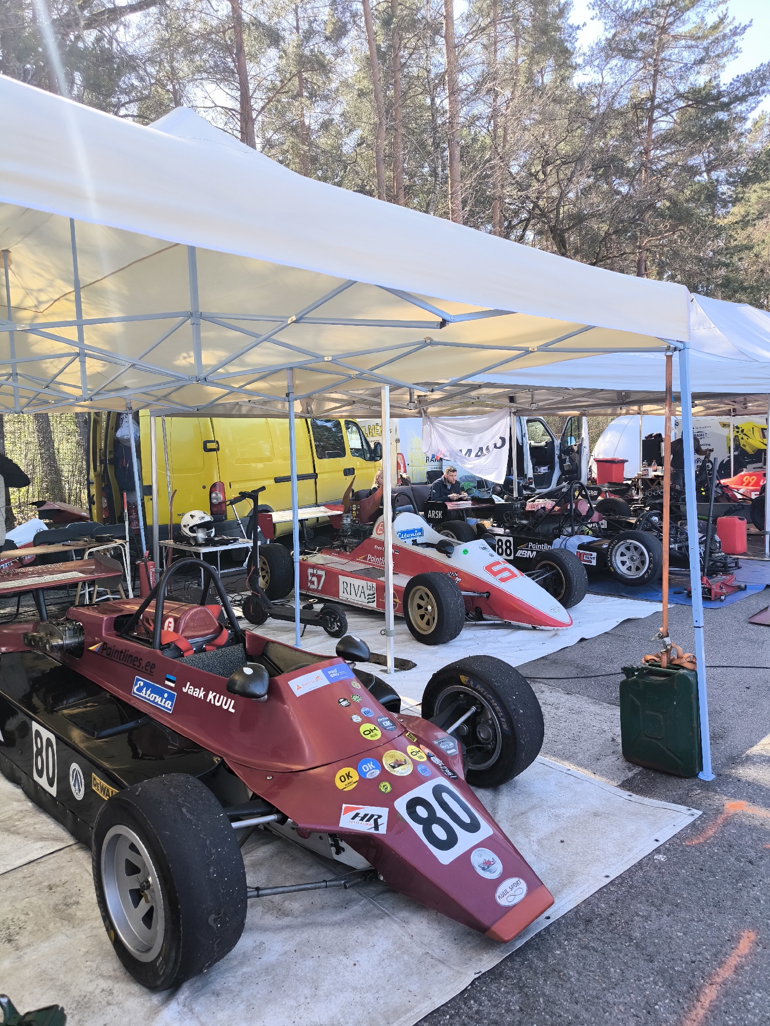 The image shows a pit area, likely at a racetrack, with several open-wheel race cars under white canopy tents. In the foreground is a maroon and black race car with the number 80 prominently displayed. Adjacent to it, further back, is another race car painted red and white, bearing the number 57. Behind these vehicles, a yellow van is visible. Farther back, additional race cars and support vehicles occupy the space under the canopies, where one individual can be seen sitting at a table. The environment appears to be a paved area with trees in the background. The image features various objects such as a gas can, tires, and equipment typical of a race team’s pit setup. The cars have sponsorship stickers and the Estonian flag, suggesting an Estonian connection, however, there's no information suggesting Riga, Latvia as a specific location.