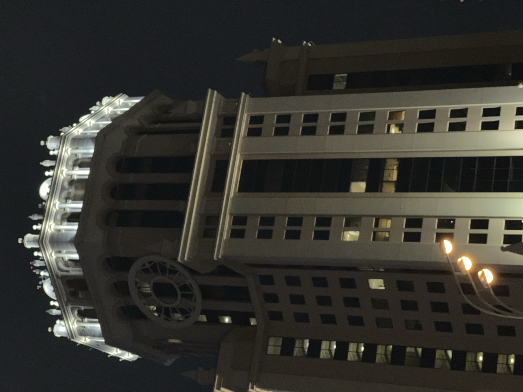 A low-angle, nighttime shot of a building in Moscow, Russia. The building has a distinct architectural style, blending classical elements with a modern facade. The left side is adorned with rounded arches, white illumination, and decorative embellishments, including a large circular window or design element. The right side has a more conventional high-rise structure with rows of rectangular windows. The lights from street lamps illuminate the building's lower sections, casting shadows and emphasizing its three-dimensional qualities. The night sky serves as the background.